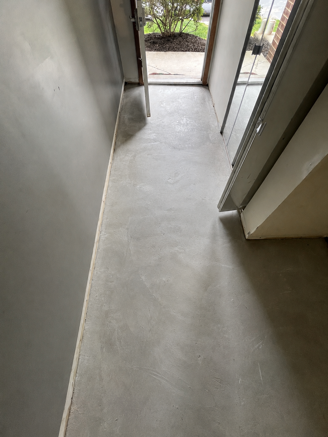 Interior view of a small entryway with a concrete floor, partially open door leading outside to a garden area, and a sliding glass door.