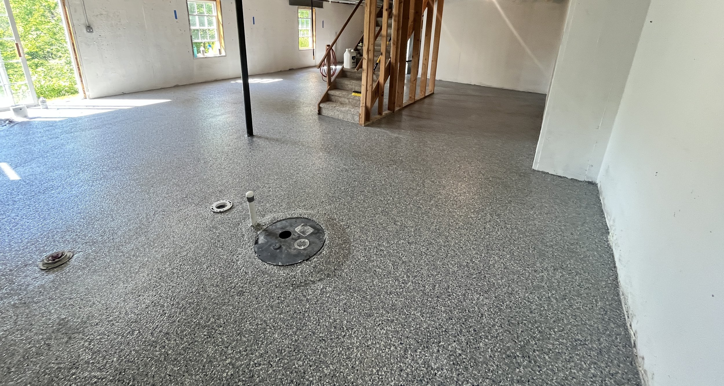 Interior of a house under construction with a newly installed speckled gray flooring, open windows allowing natural light, and a staircase with exposed wooden framing.