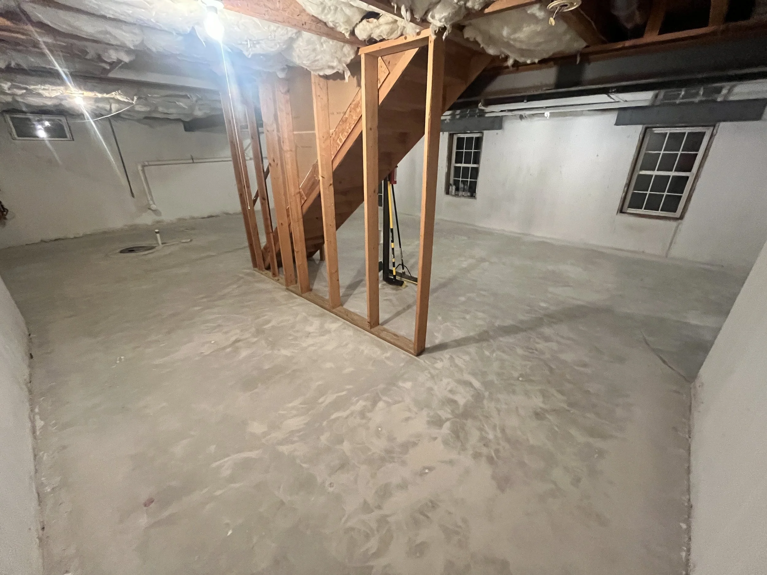 Unfinished basement with concrete floor, exposed insulation and ceiling, small staircase with wooden framing, two small windows.