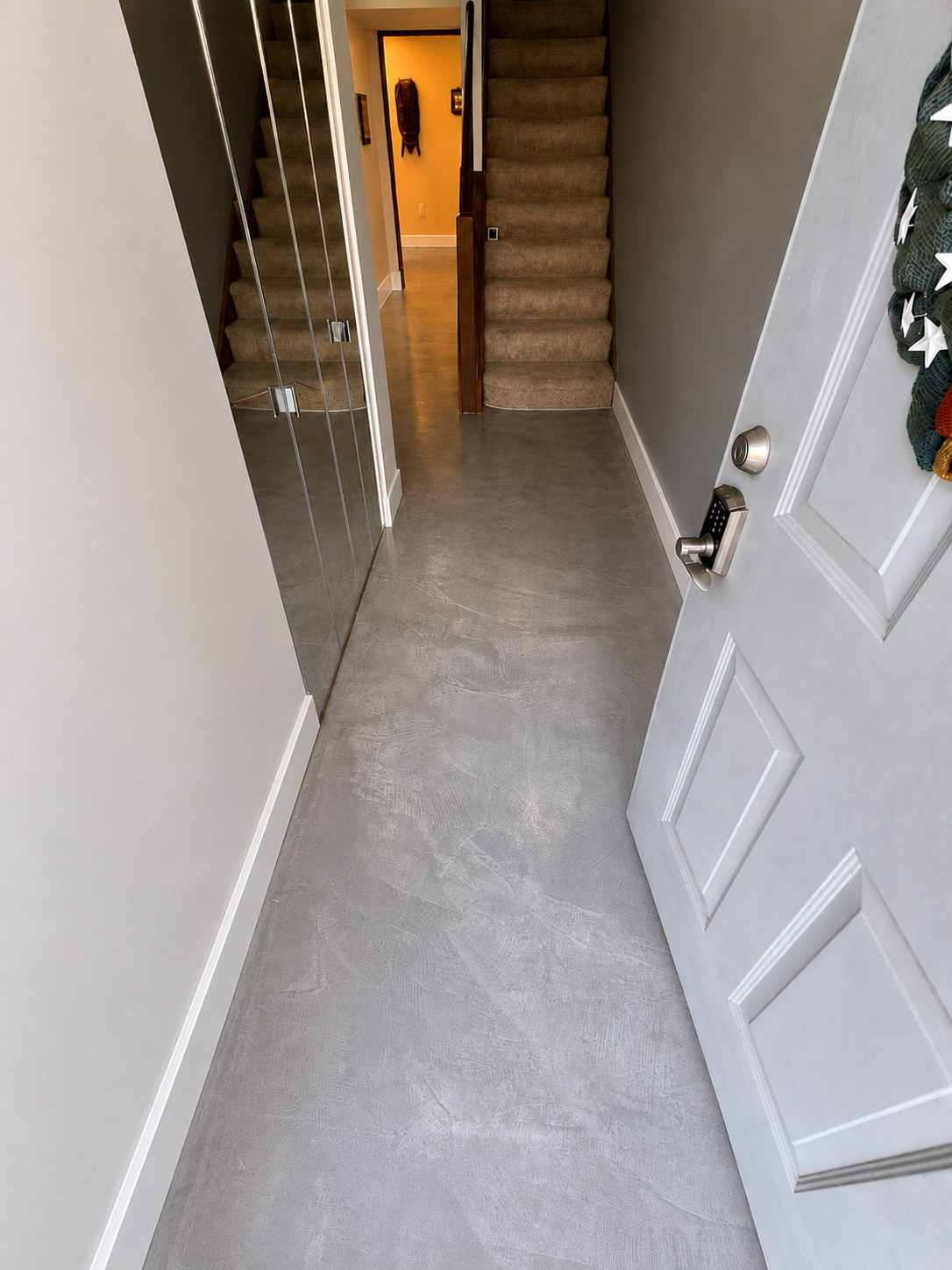 Entryway with a white door, a digital lock, a wreath hanging on the door, mirror closet doors on the left, wood staircase with carpeted steps ahead, and a hallway leading to a warmly lit room.