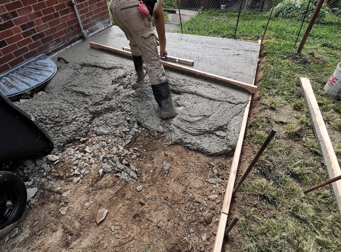 A person is pouring and leveling concrete in a framed area next to a brick house with grass around.