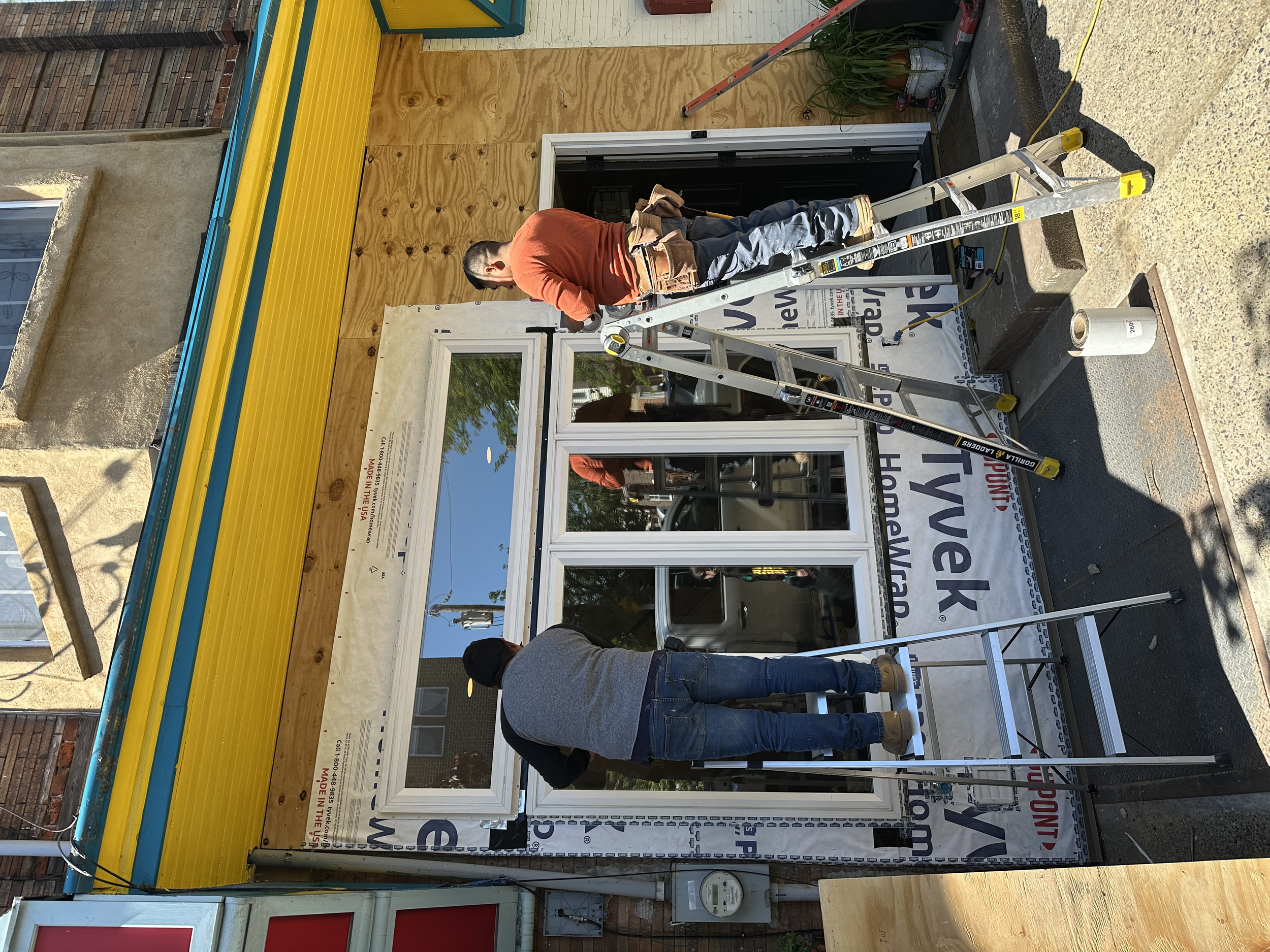A man working on the installation of a storefront window, standing on a ladder in front of a building with plywood sheathing and a partially covered door. The scene appears to be a construction or renovation site.