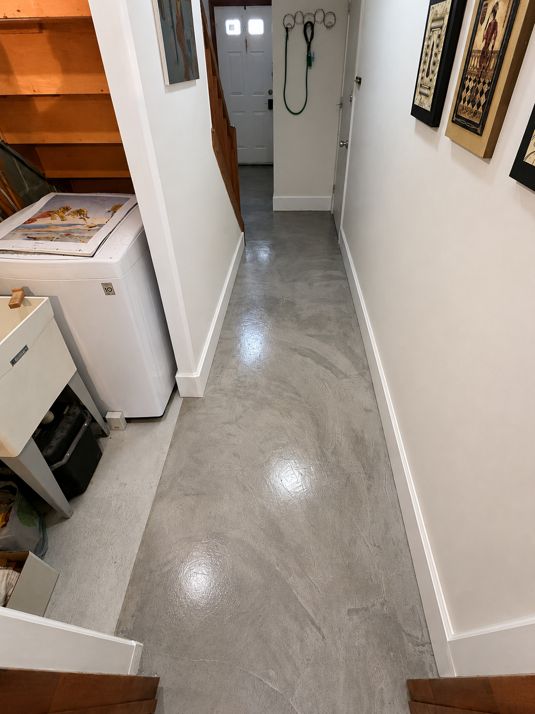 A narrow laundry room with polished concrete flooring, white walls, and a white washing machine on the left. There are framed artworks on the right wall, hooks on the wall near the door, and a staircase leading up on the left side. The front door is 