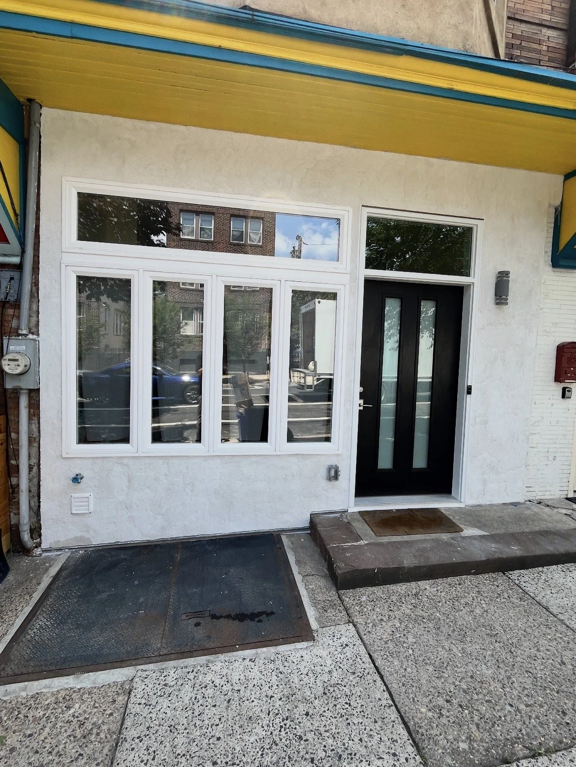 The image shows a modern storefront entrance with white framed windows and a black door set into a white wall. There is a small step and a welcome mat outside. The area features a concrete sidewalk with a metal ramp, and neighboring buildings are vis