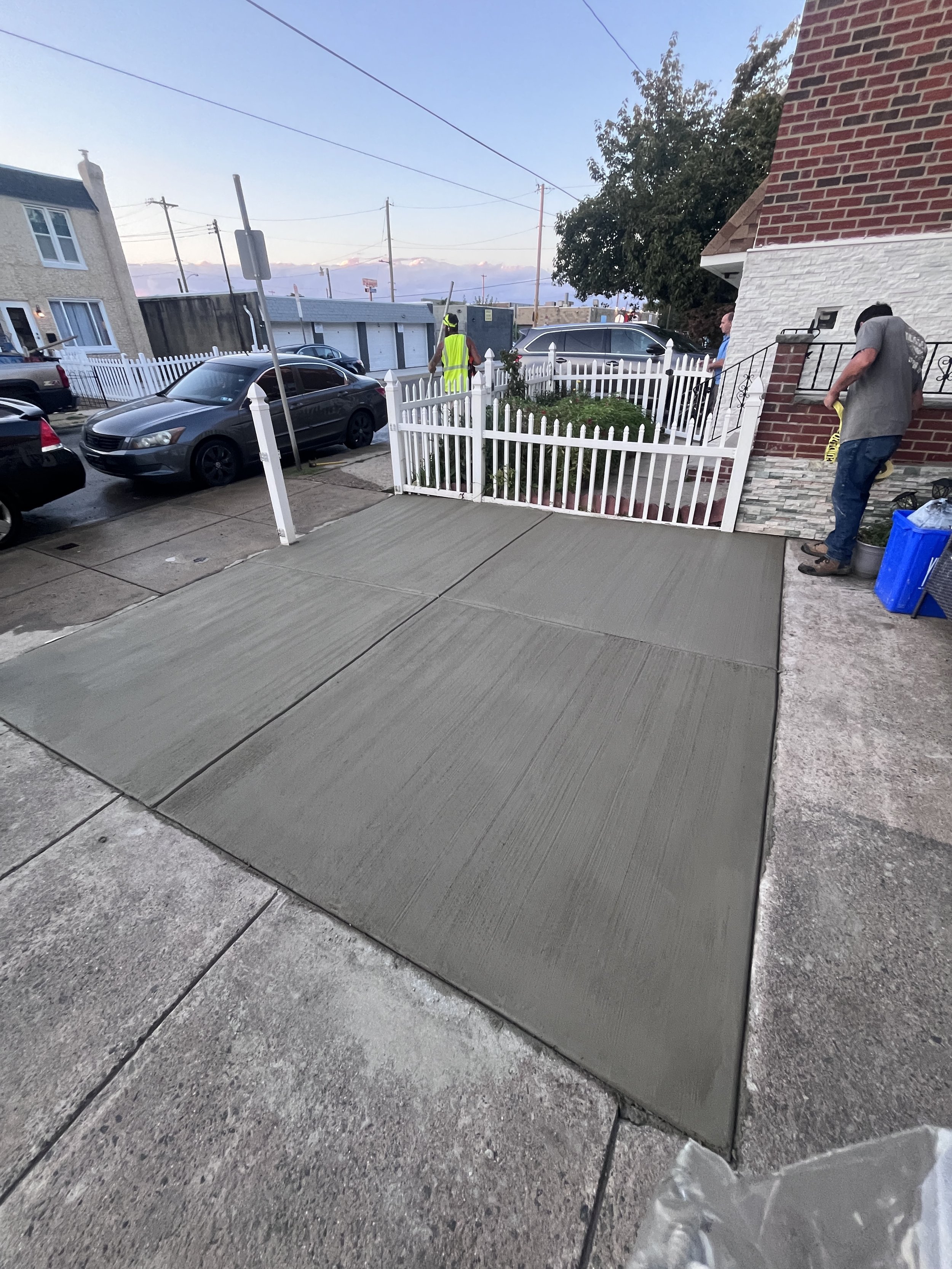Newly poured concrete sidewalk in front of a house with a brick exterior, white picket fence, and parked cars. Three men are present, one in a high-visibility vest working near the fence and two others, one in a gray shirt and the other in a blue shi