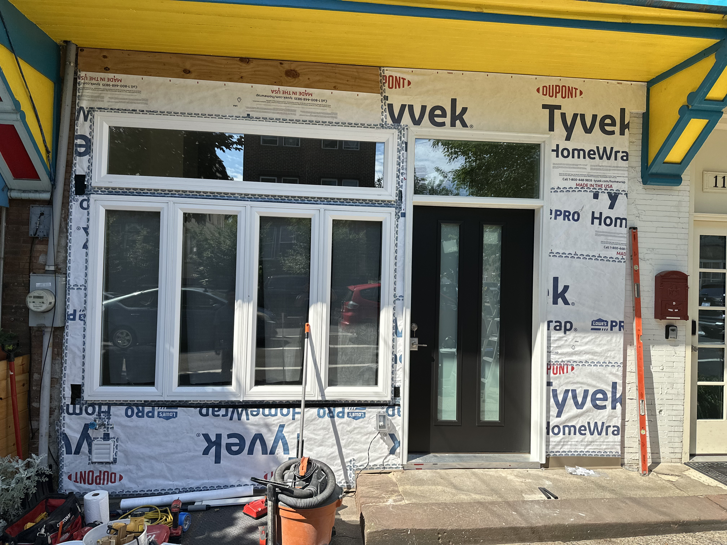 Front of a house under construction with window and door frames installed, covered in Tyvek HomeWra weather barrier, construction tools and materials in the foreground, and partially finished yellow and teal exterior siding.