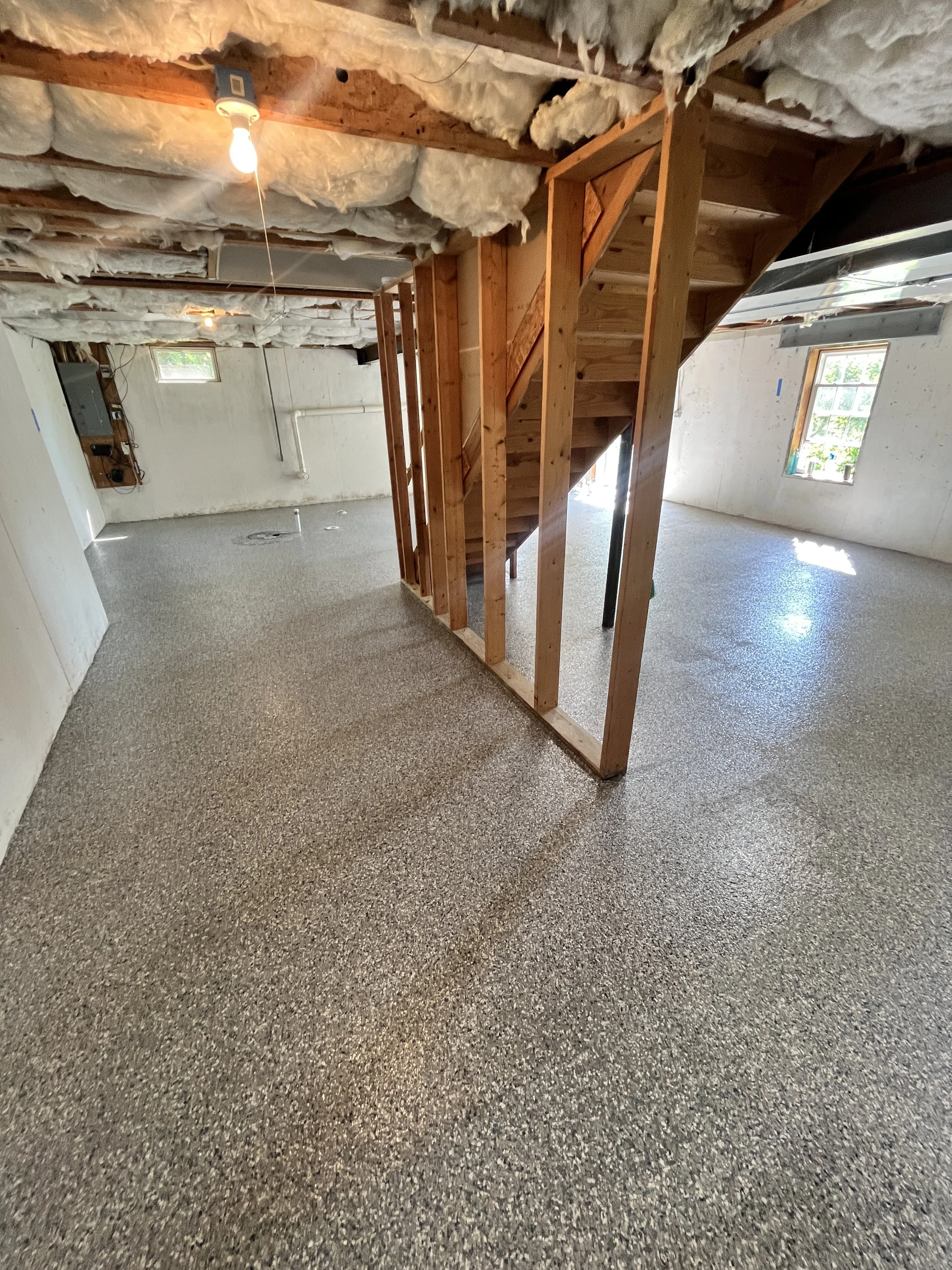 Interior of a basement under construction with exposed wooden framing, insulation on the ceiling, and new flooring. There are small windows letting in natural light.
