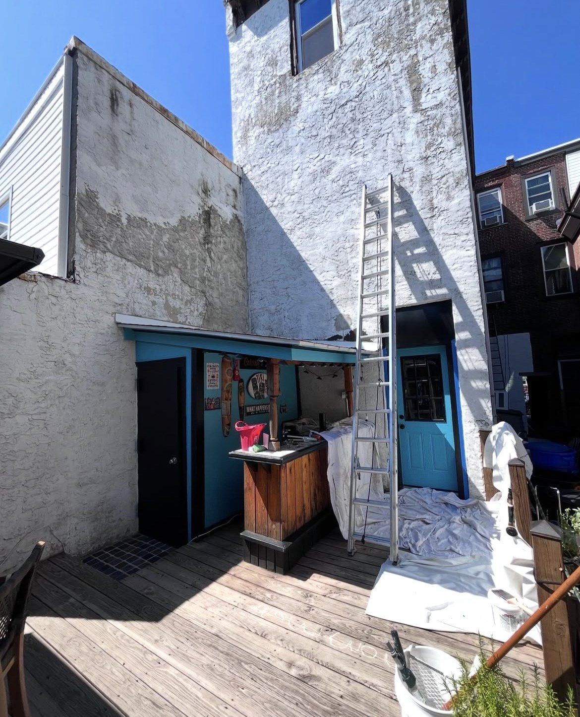 Backyard patio with wooden deck, a ladder leaning against a white textured building, a blue door, a small bar area with a counter, and a tarp-covered area under construction. Sunny day with clear blue sky.