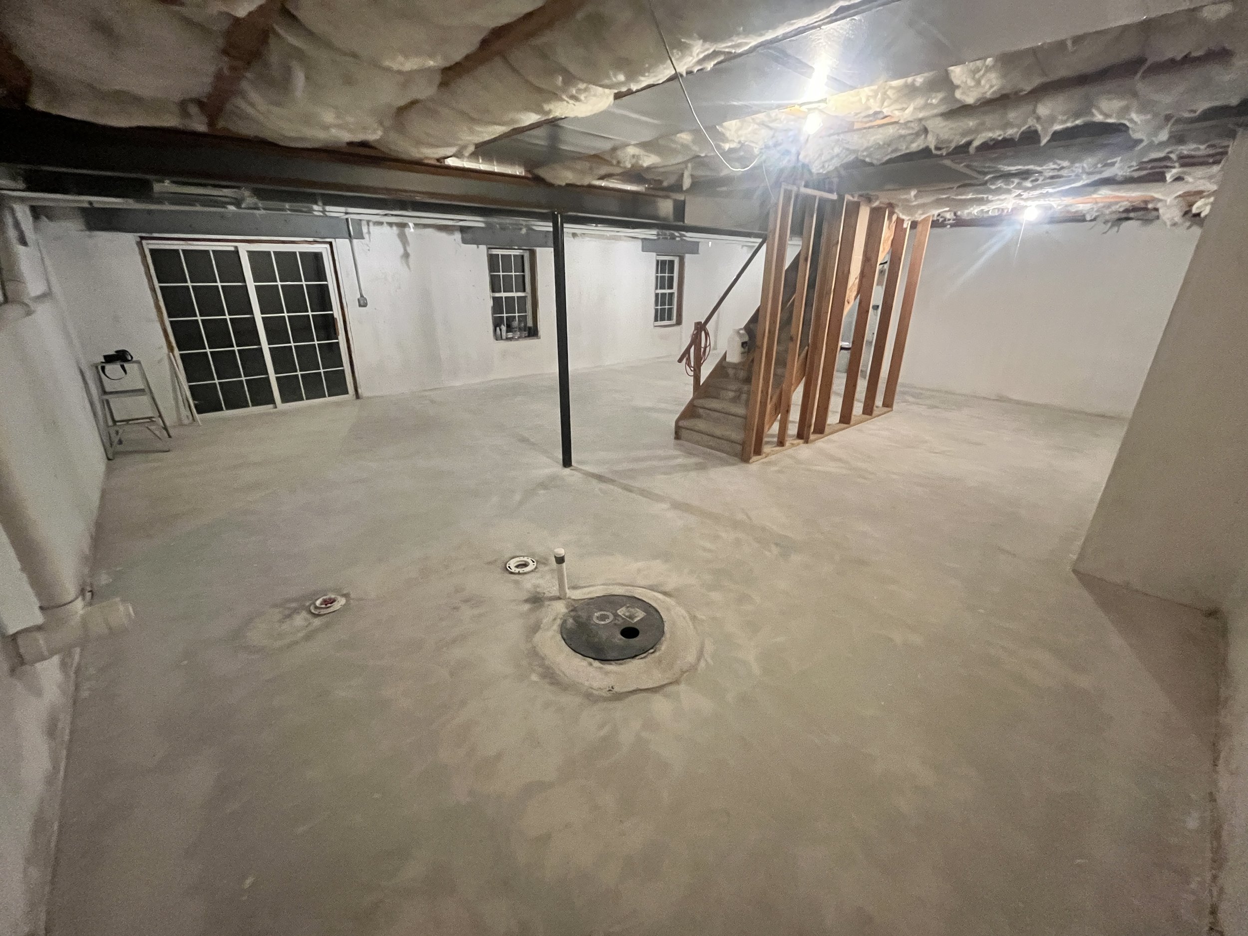 Basement room under construction with unfinished walls, exposed insulation on the ceiling, a staircase on the right, and plumbing pipes on the floor.