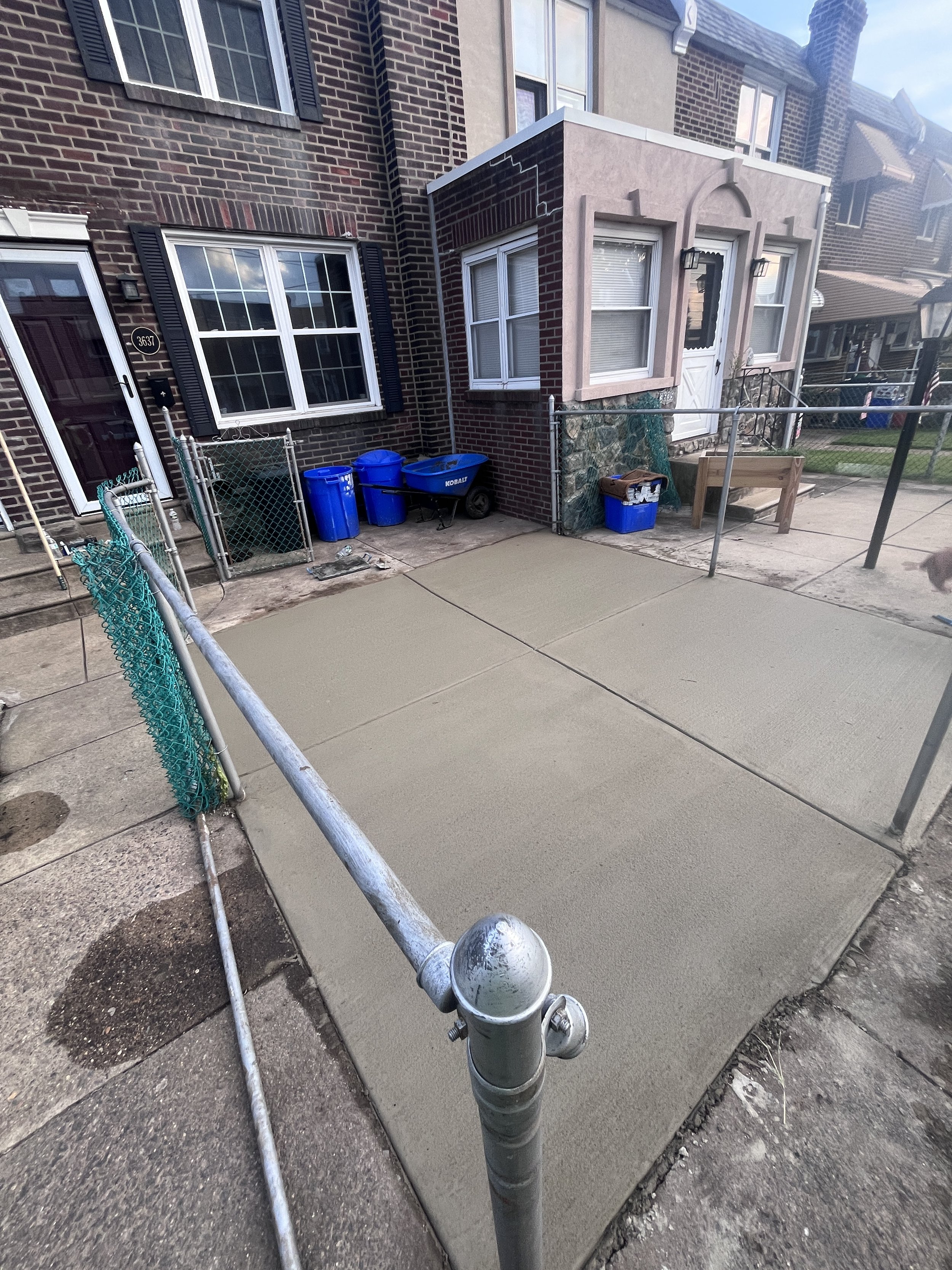 View of backyard patio area with freshly poured concrete, fenced with metal fence, with bricks and siding house in the background, and construction materials and blue recycling bins nearby.