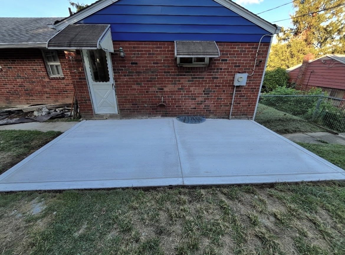 Backyard with a newly poured concrete patio, brick house wall with small window and air conditioning unit, grass area surrounding the patio, and neighboring house with fencing.