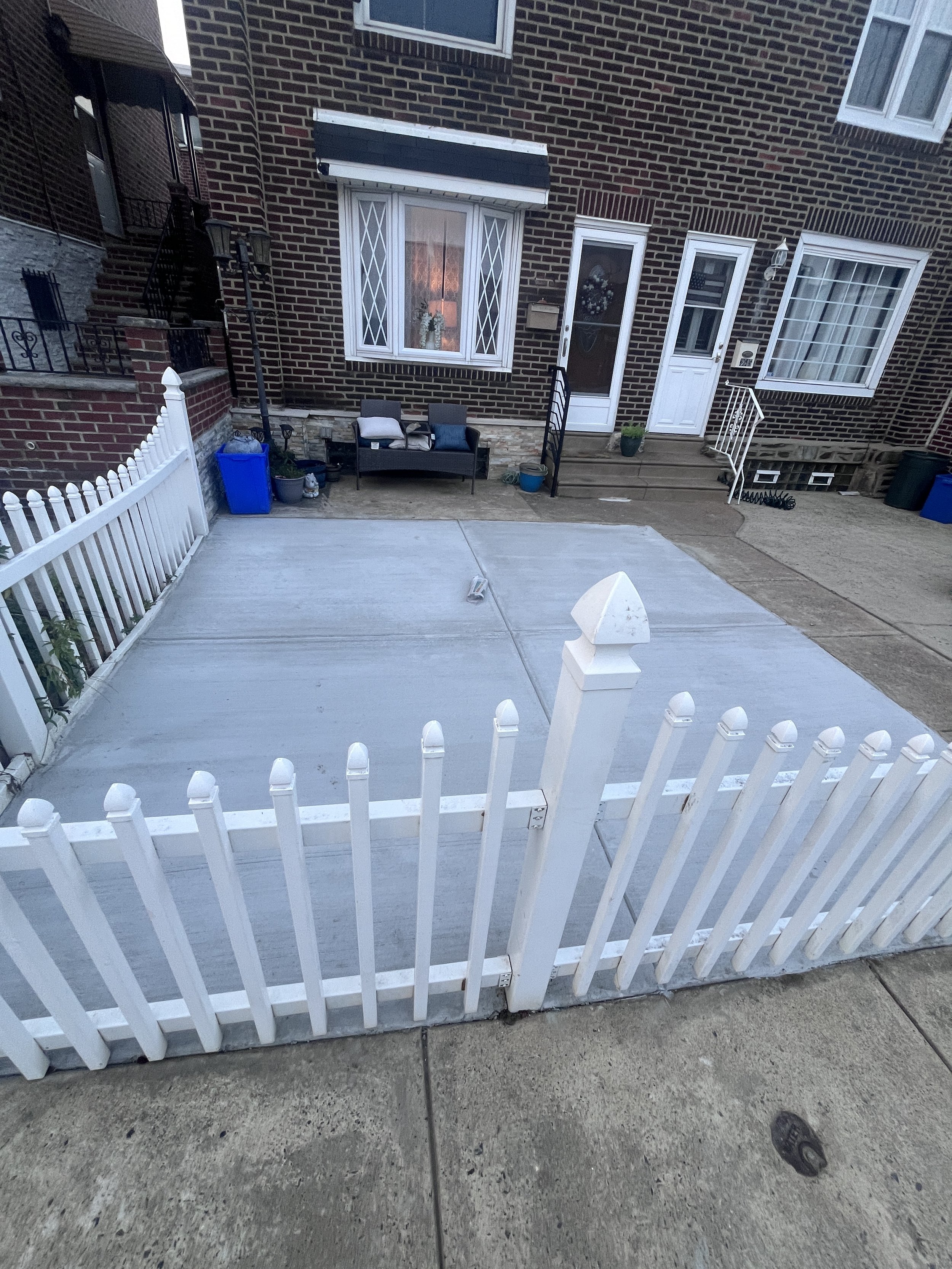 View of a residential backyard patio with a white picket fence, concrete surface, and brick house in the background. There is outdoor furniture, potted plants, and a trash can. The patio is surrounded by a brick wall and steps leading to the house en