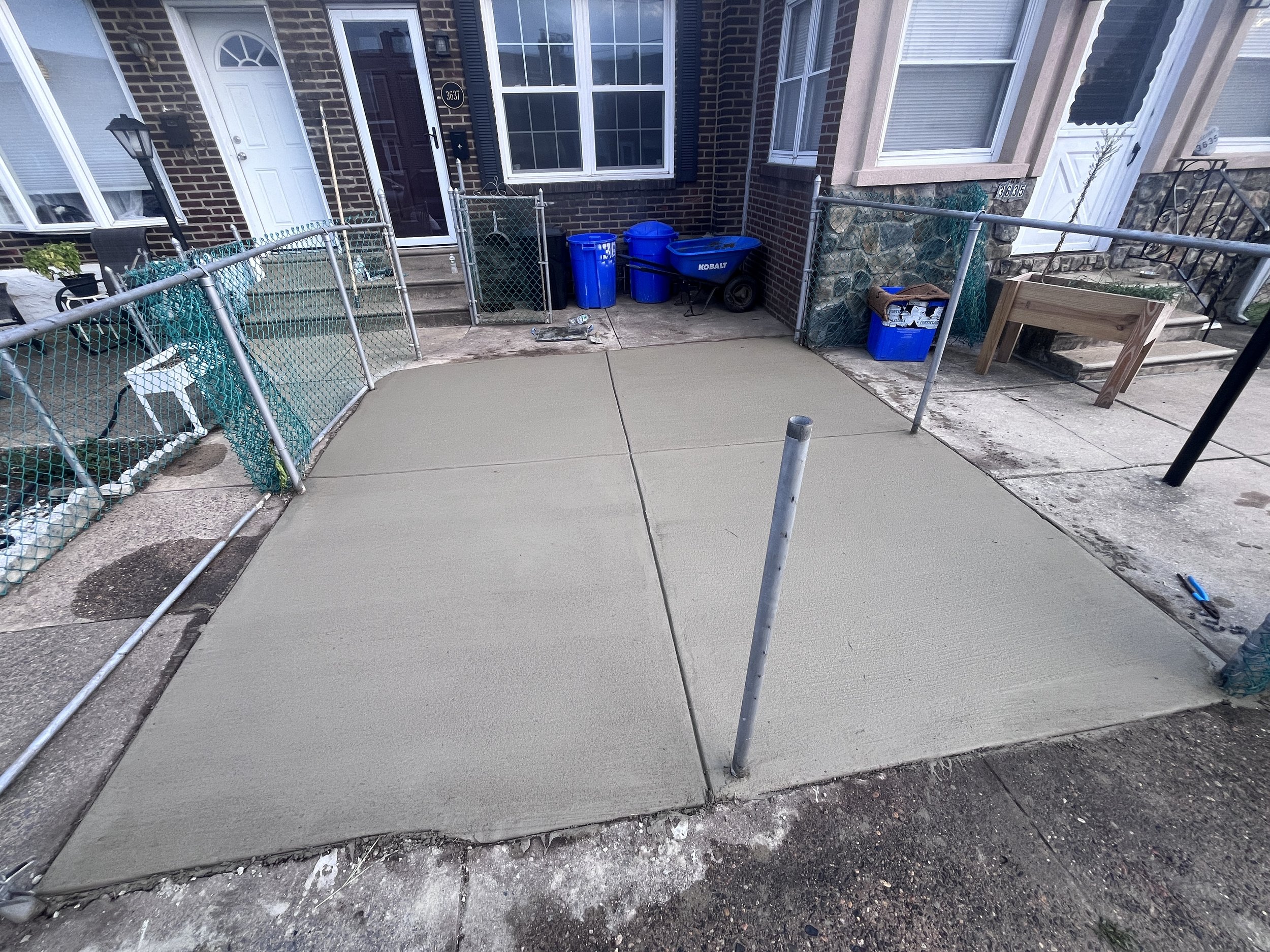 Newly poured concrete driveway in front of a brick residential building with stairs, windows, and various garbage and recycling bins.