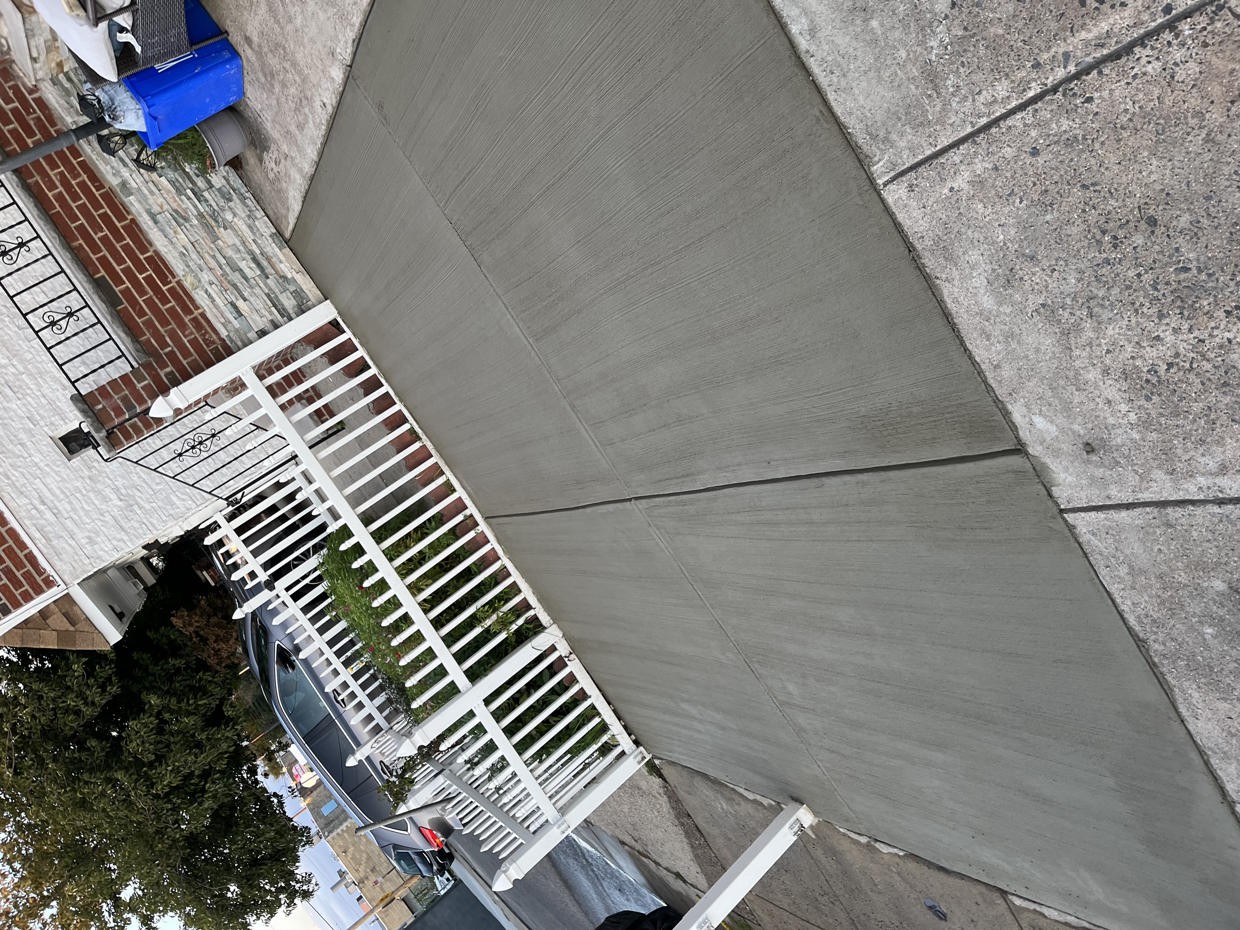 A sidewalk with a curved section of freshly poured concrete next to a brick building with a white fence. Vehicles are parked in the background, and there are trees and streetlights.