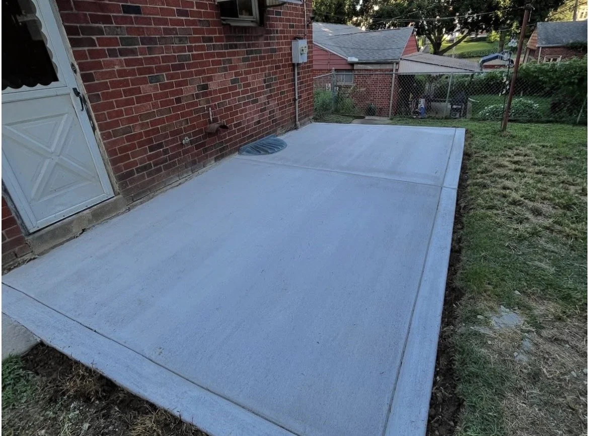 Freshly poured concrete patio next to a red brick house with a white door, a vent, and outdoor electrical box, enclosed by a chain-link fence with neighboring houses nearby.