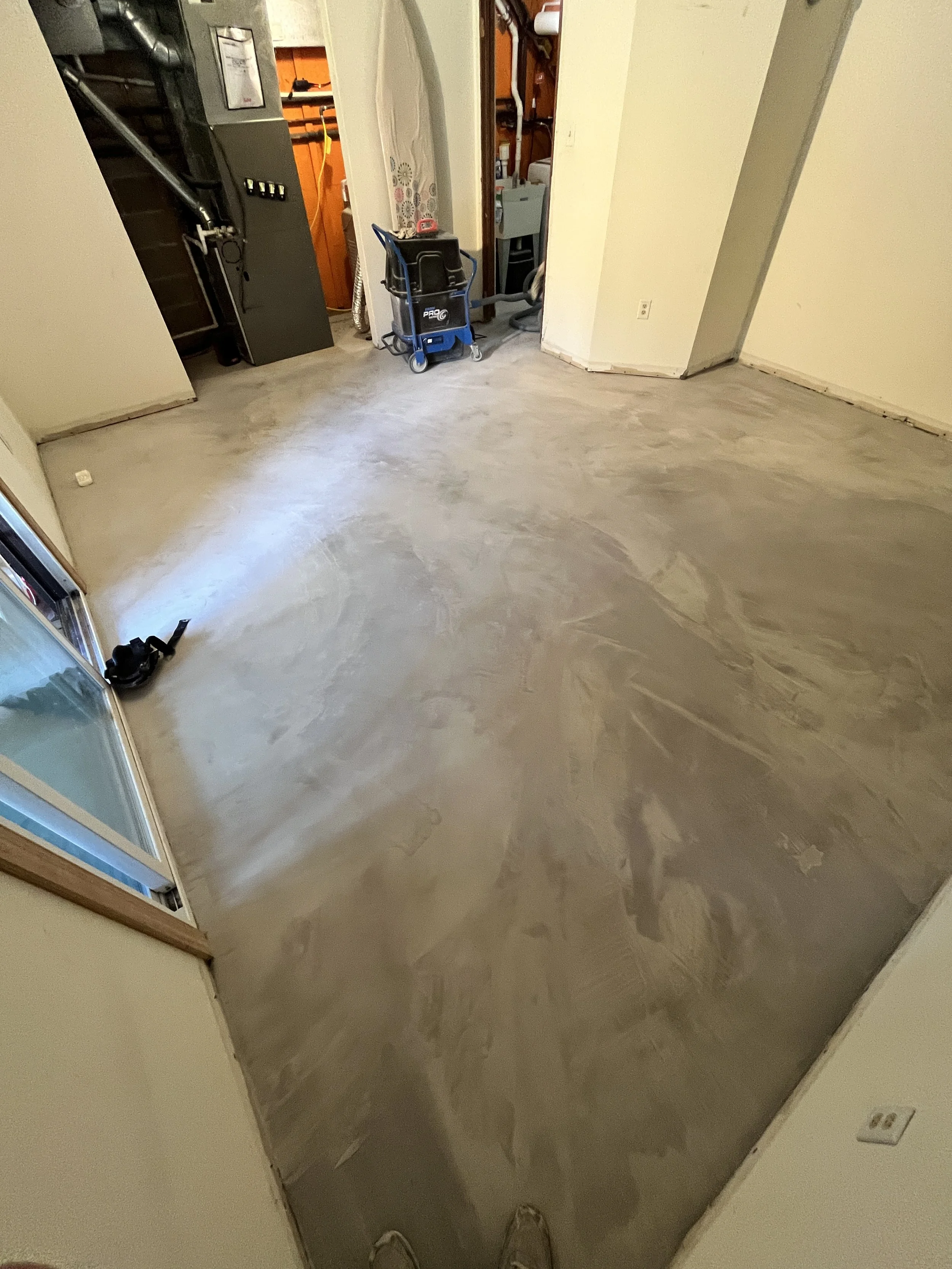 Empty room with a concrete floor, a small window, and a doorway leading to a storage or utility area with a vacuum cleaner and various tools.