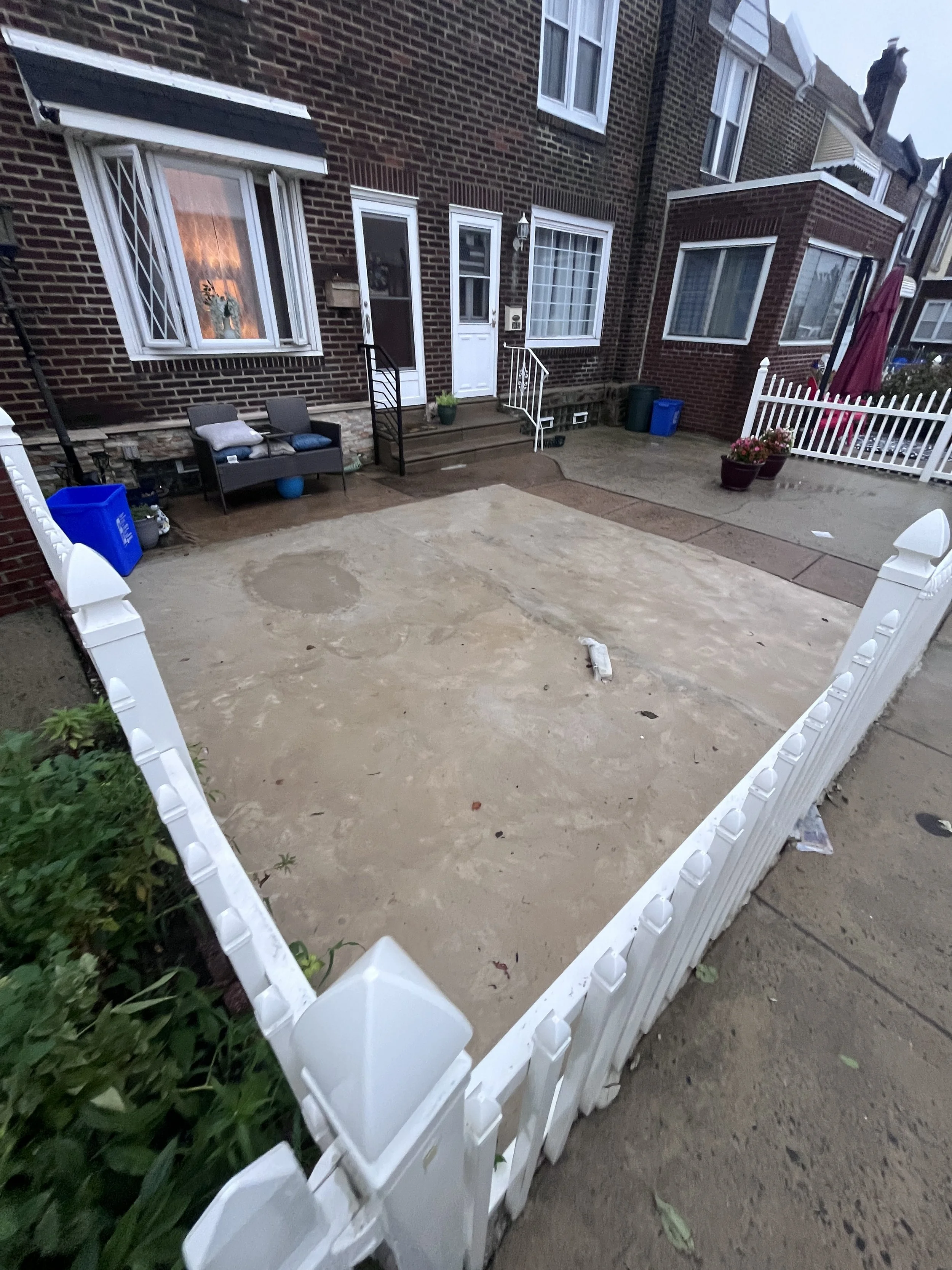 Front yard of a brick house with a white picket fence, potted plants, outdoor chairs, and a concrete patio.