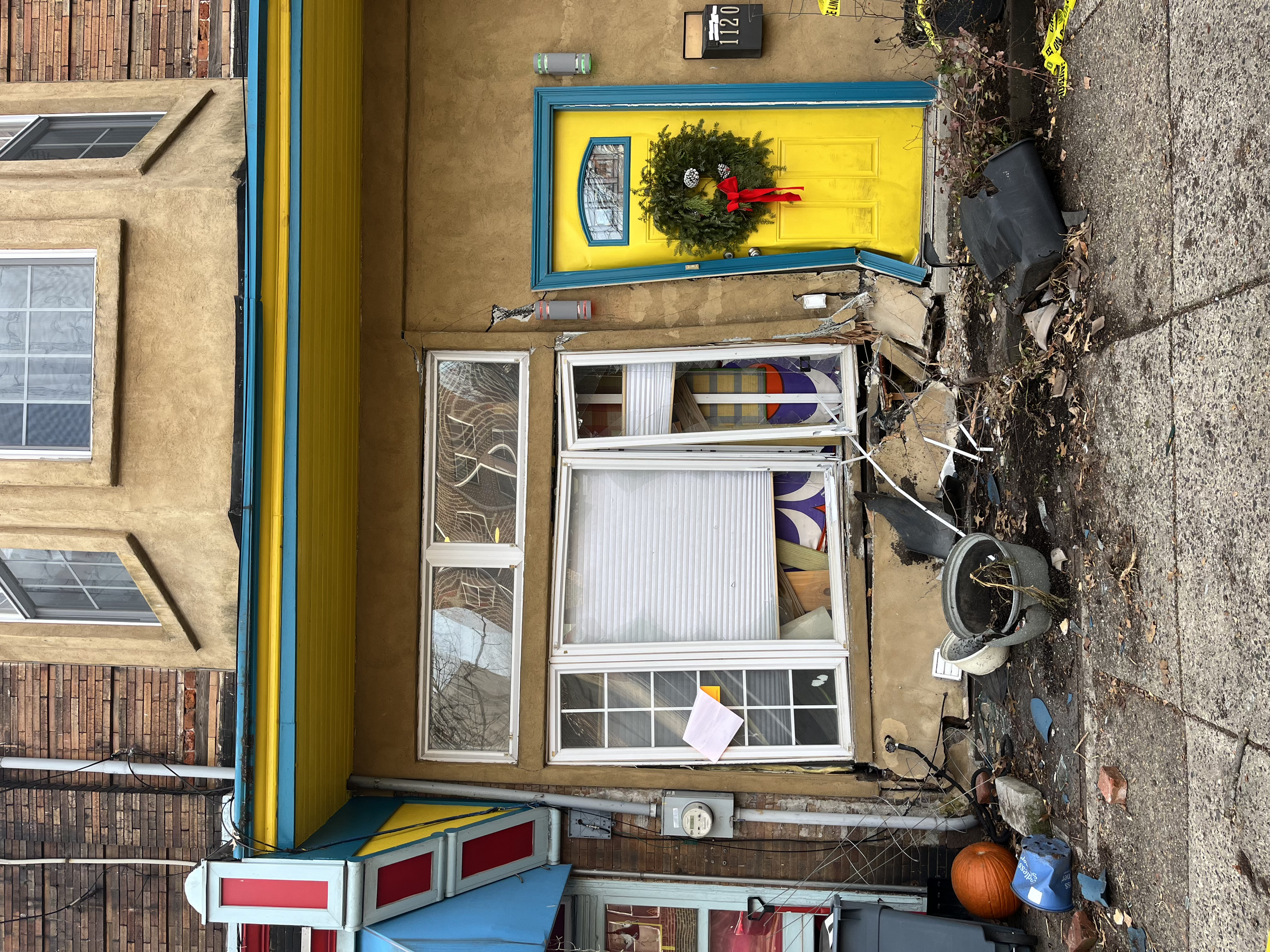 Front of a building with a yellow door decorated for Christmas with a wreath and red bow. The building has tan stucco walls, windows with white frames, and a blue and yellow striped awning. There is construction debris and a pumpkin outside in a blac