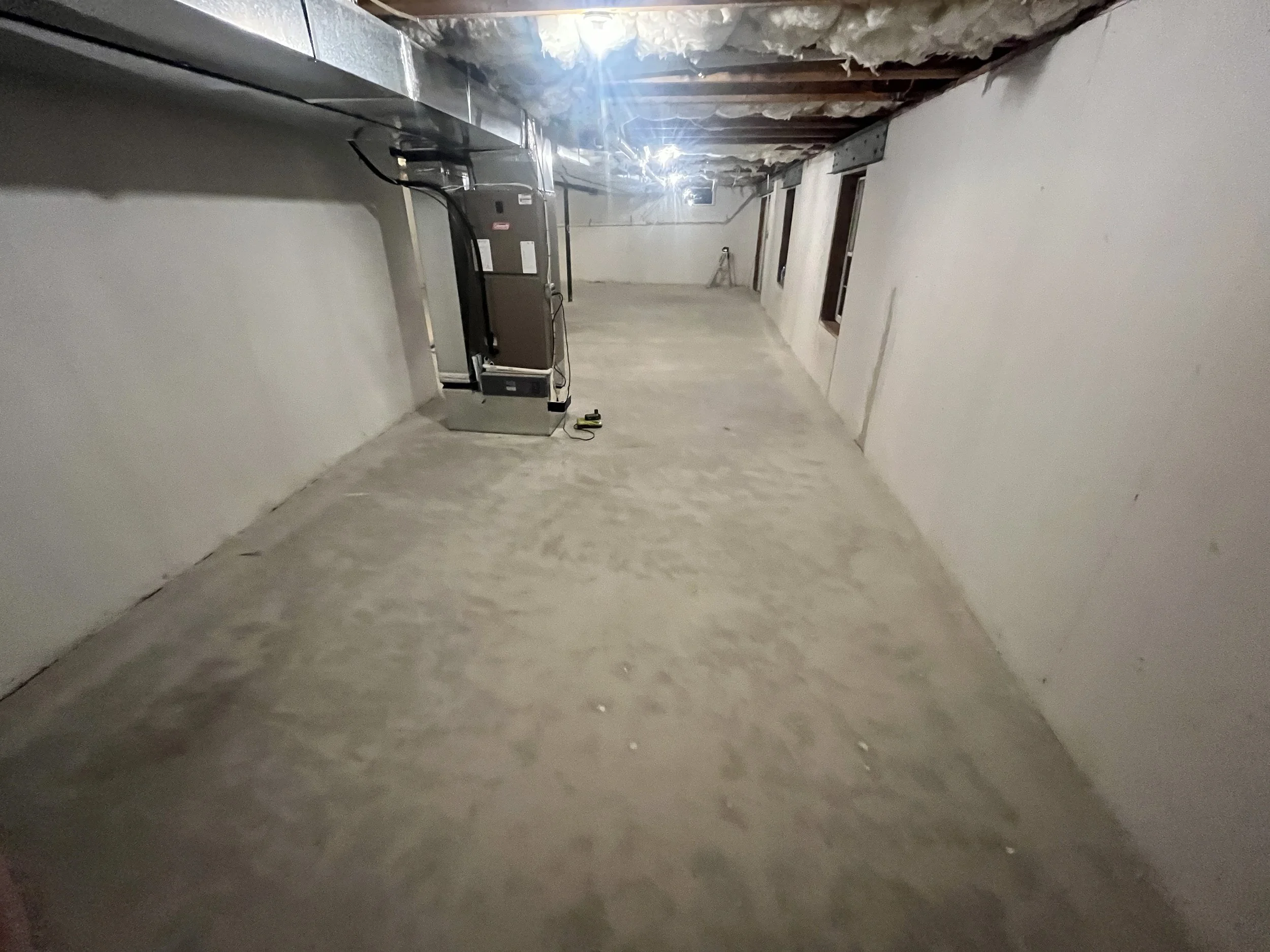 A basement or utility room with unfinished ceiling, white walls, and a concrete floor. There is a furnace or HVAC unit and some tools on the floor, with exposed ceiling joists and insulation visible at the top.
