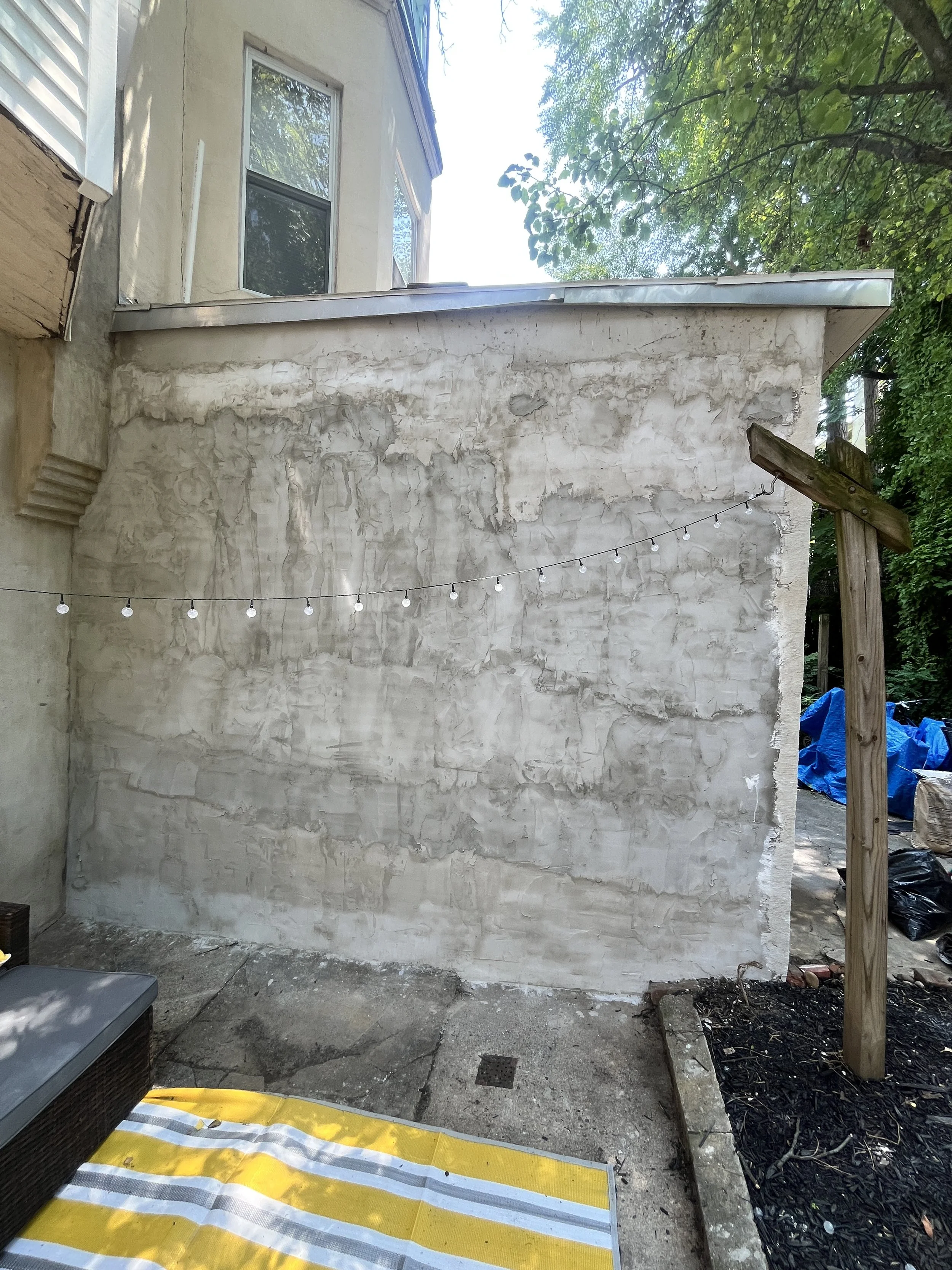 Backyard scene showing a partially renovated wall with exposed stucco and a string of outdoor lights, surrounded by green trees and patio furniture.