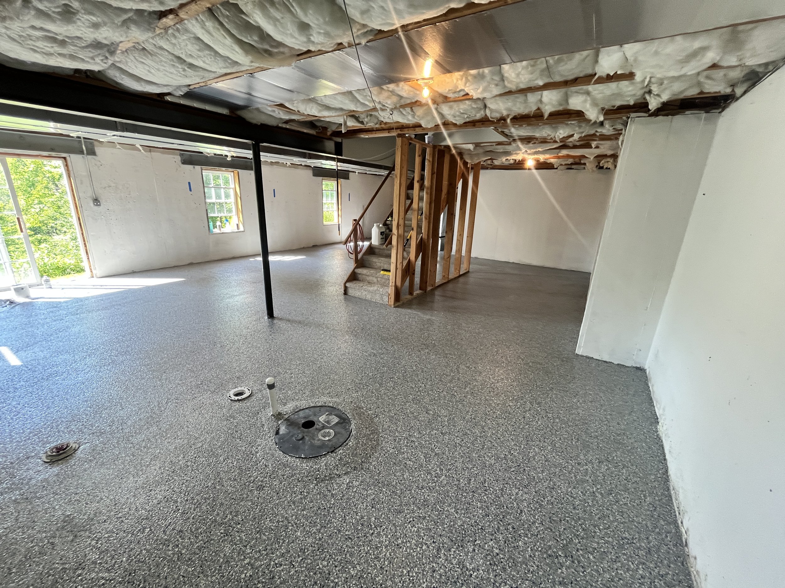Construction in progress inside a house with insulated ceiling and unfinished walls. The floor is prepared for plumbing with circular cutouts, and a wooden staircase is built leading to an upper level. There are two windows letting in natural light.
