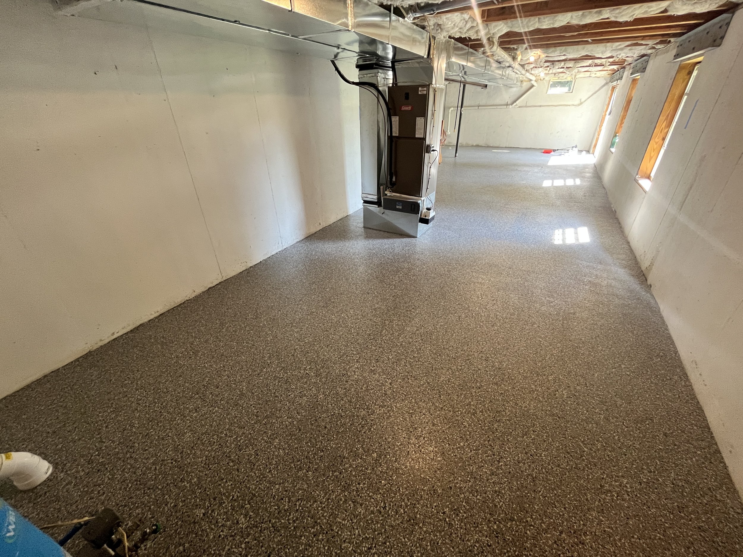An unfinished basement with carpeted flooring, exposed ceiling beams, ducts, and insulation. There are three small windows allowing natural light. Construction tools and equipment are visible, including a furnace and a shoe or boot for scale.