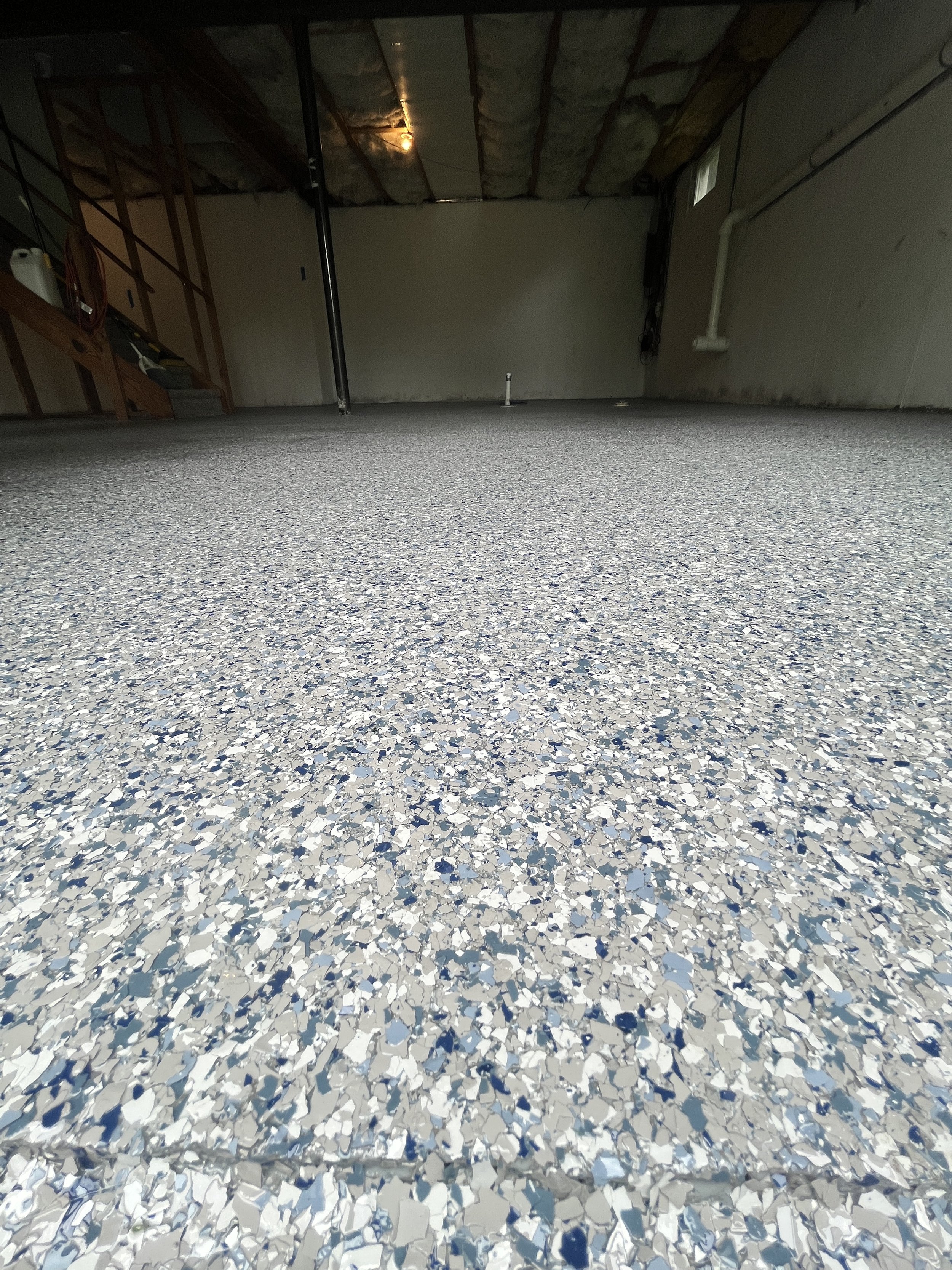 Room with a textured blue-gray epoxy floor, unfinished drywall and ceiling insulation, exposed pipes, and a staircase leading up on the left side.