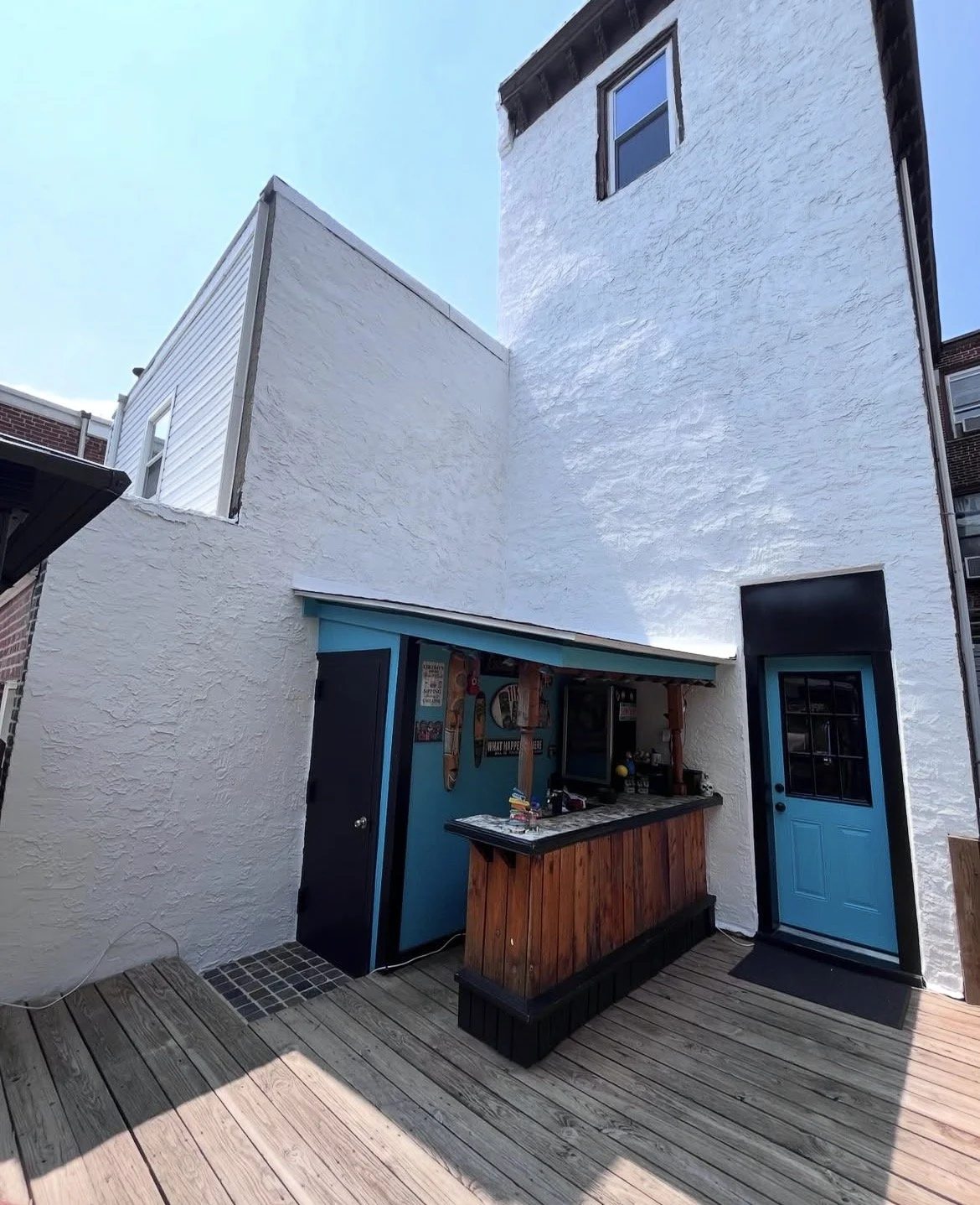 Small outdoor bar with blue walls, black countertop, and wooden paneling, situated against a white textured exterior wall of a building under a clear sky.