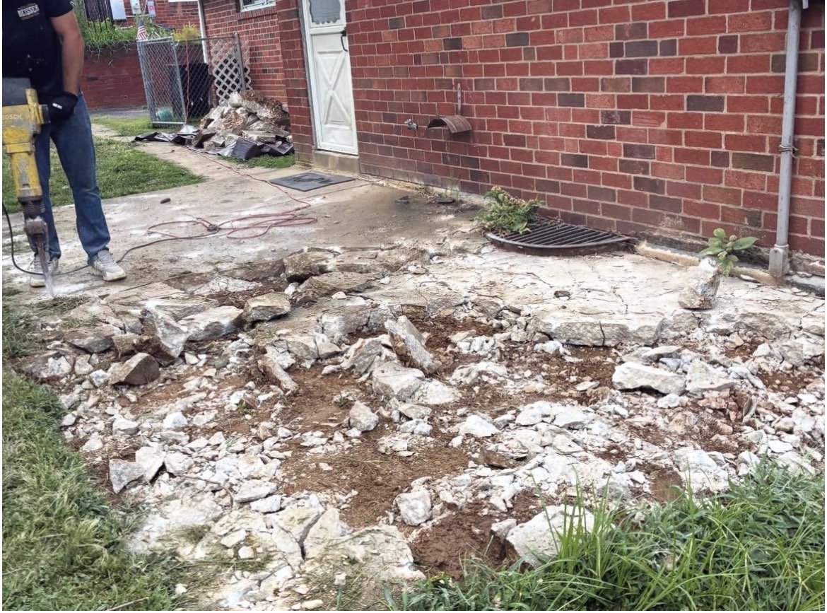 A person holding a jackhammer working on breaking up a concrete patio outside a brick house, with debris scattered around.