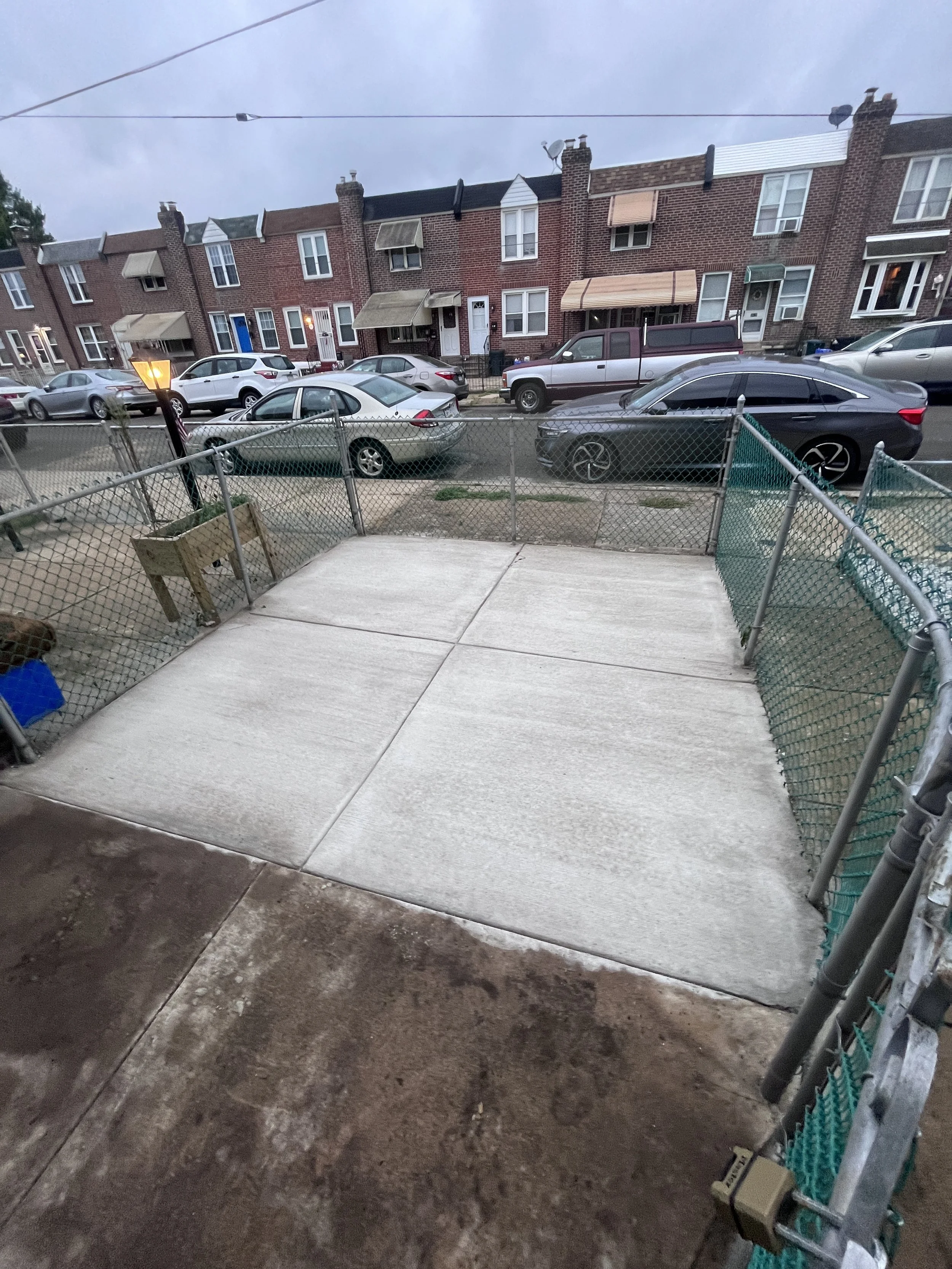 View of a residential street with brick row houses and parked cars, seen from a backyard porch with a concrete floor and metal fencing.