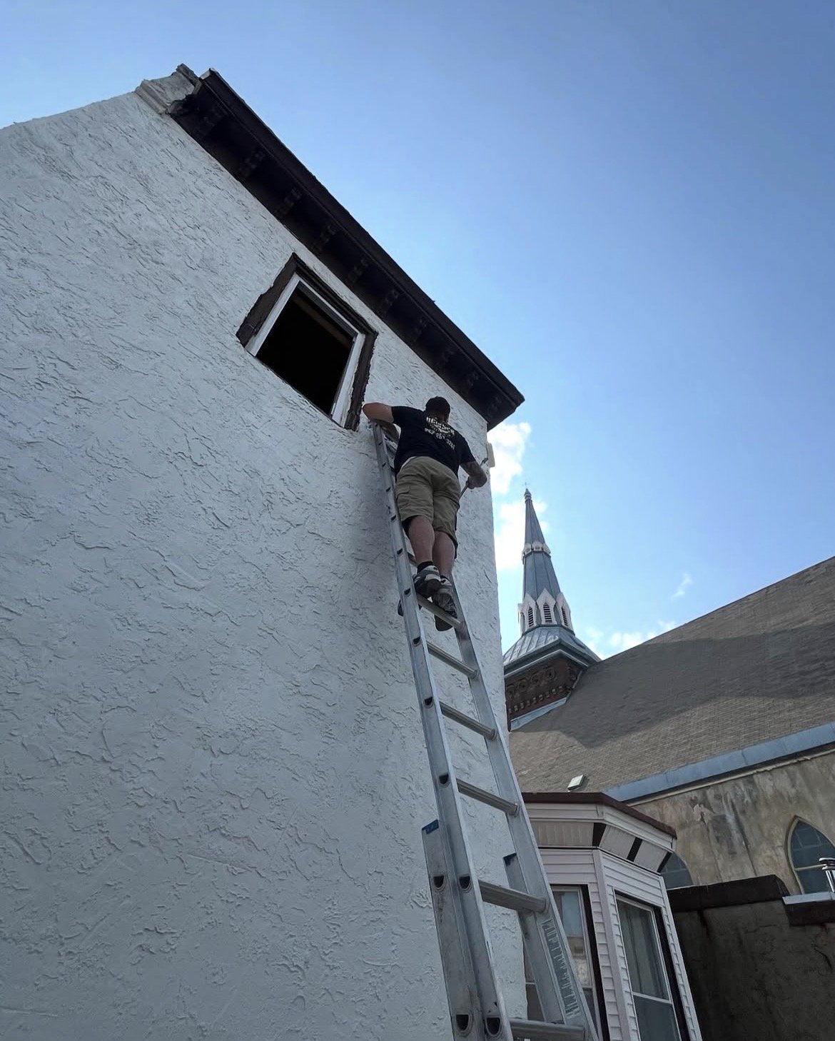 A man standing on a ladder, working on the exterior wall of a white building near a window, with a church steeple visible in the background.