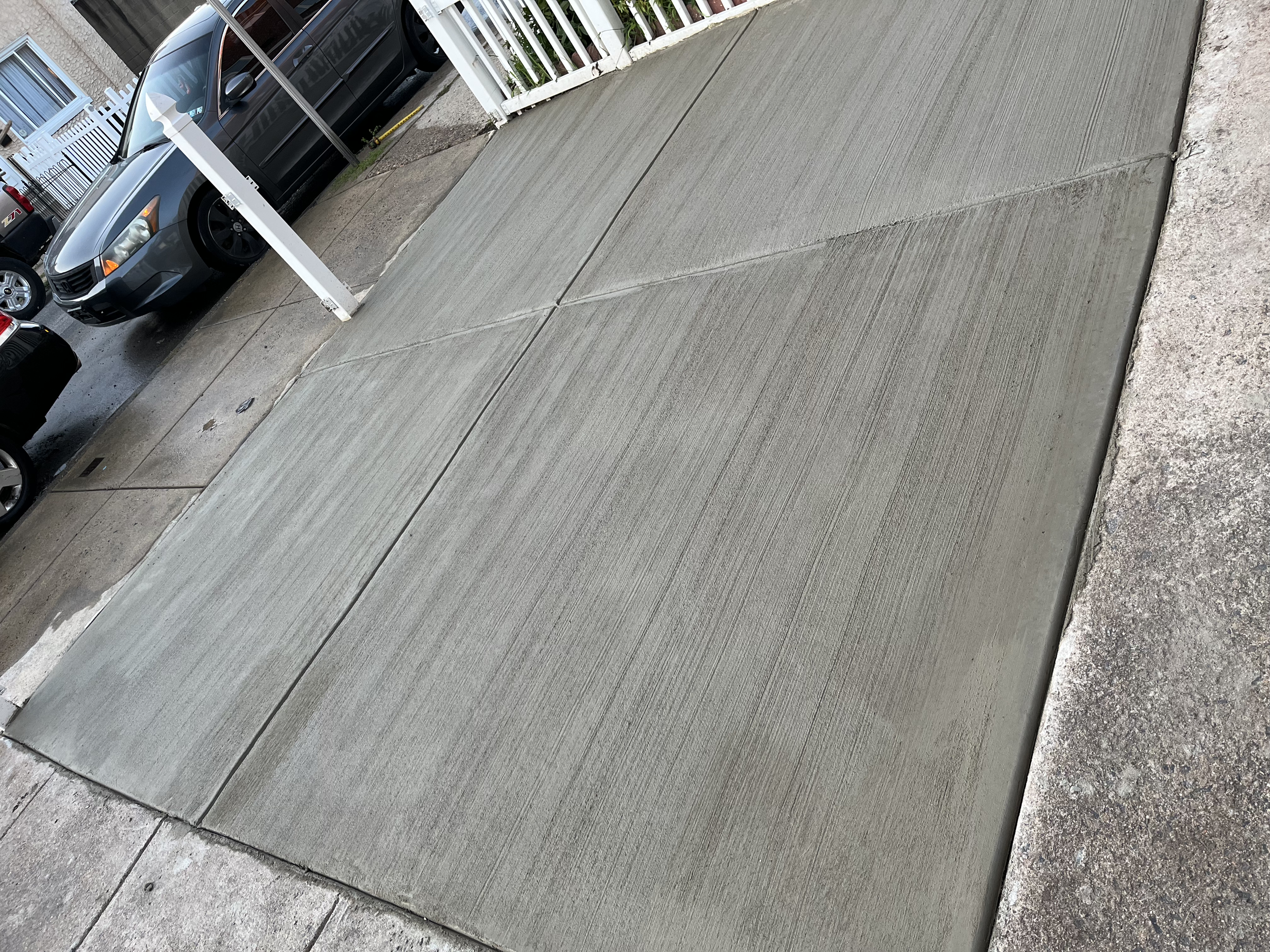 Freshly poured concrete sidewalk next to a residential driveway, with cars parked nearby and a white fence.