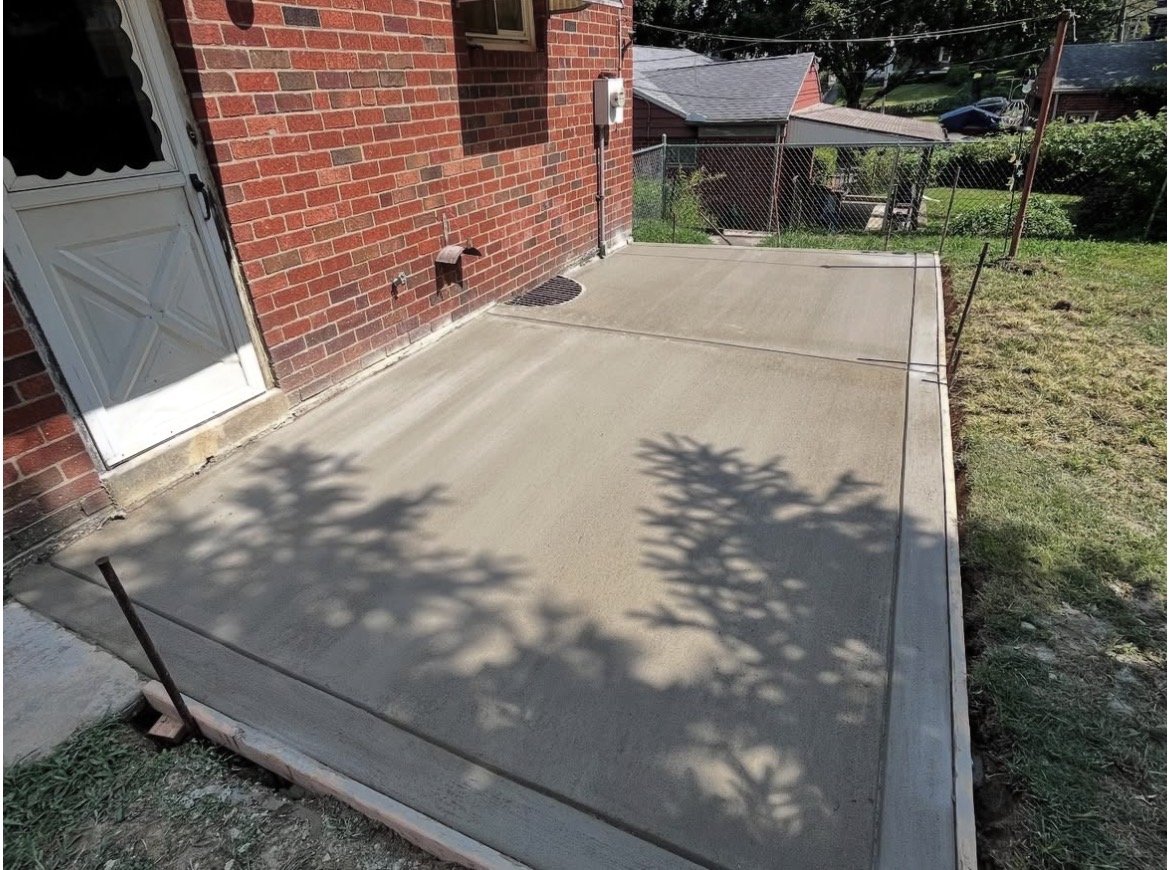 Freshly poured concrete patio with shadows of tree branches and leaves, next to a red brick house with a white door and nearby grassy yard.