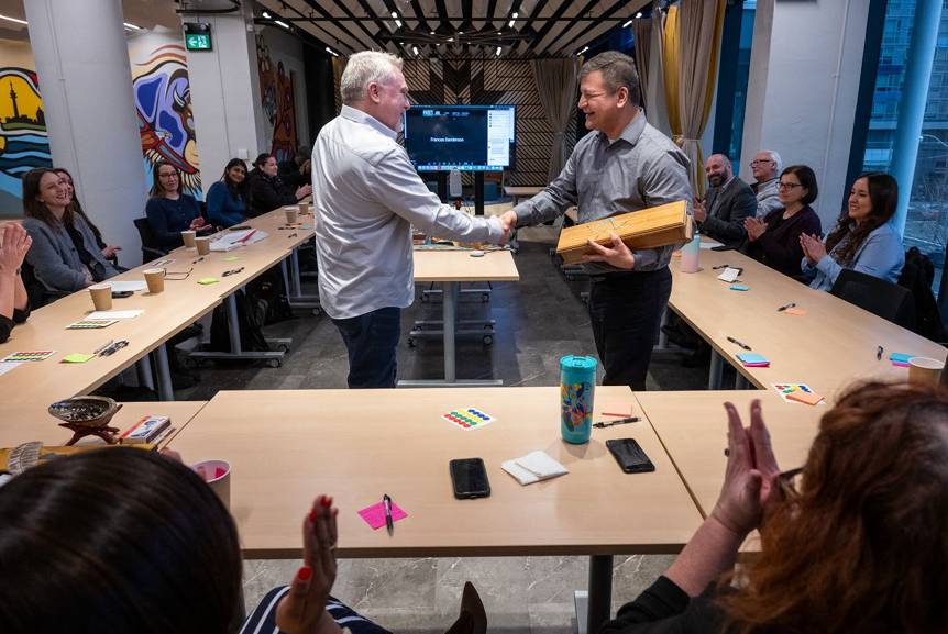 Two men shaking hands in a conference room, one of them holding a wooden box, with seated people clapping around them.