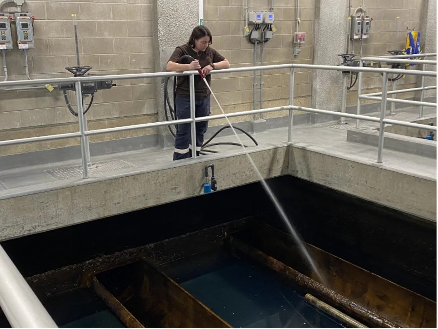 A woman wearing work clothes and gloves is cleaning a large industrial water tank using a hose, standing on a concrete platform with safety railings inside a facility with electrical equipment on the wall.