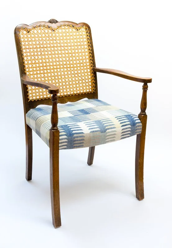 An antique wooden armchair with a woven cane backrest and upholstered seat featuring a blue and white geometric pattern.