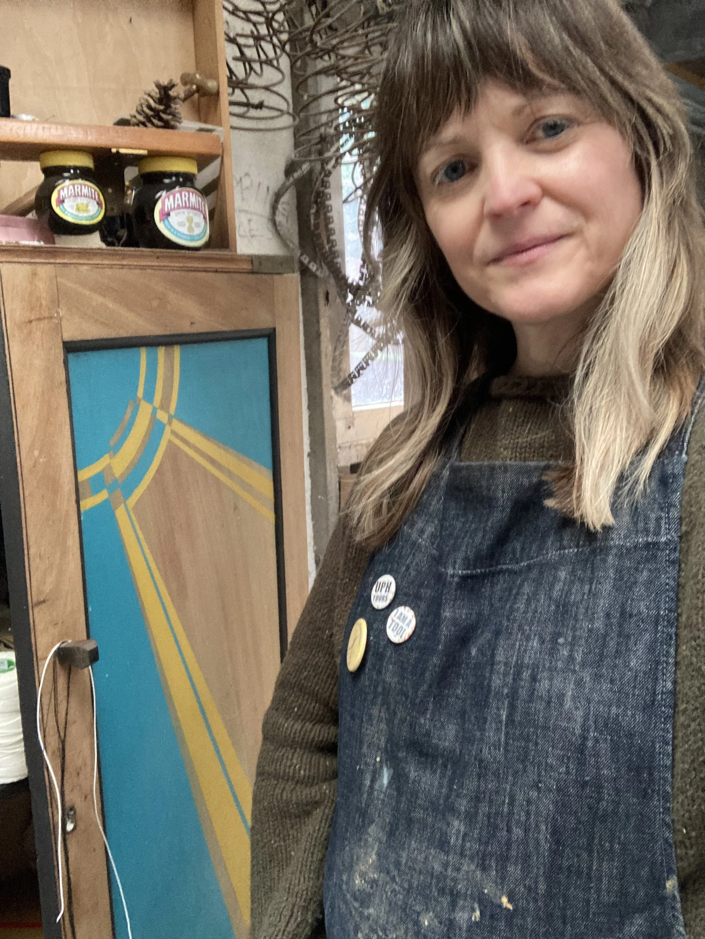 A woman with long blonde hair wearing a dark denim vest stands in front of a wooden display shelf.