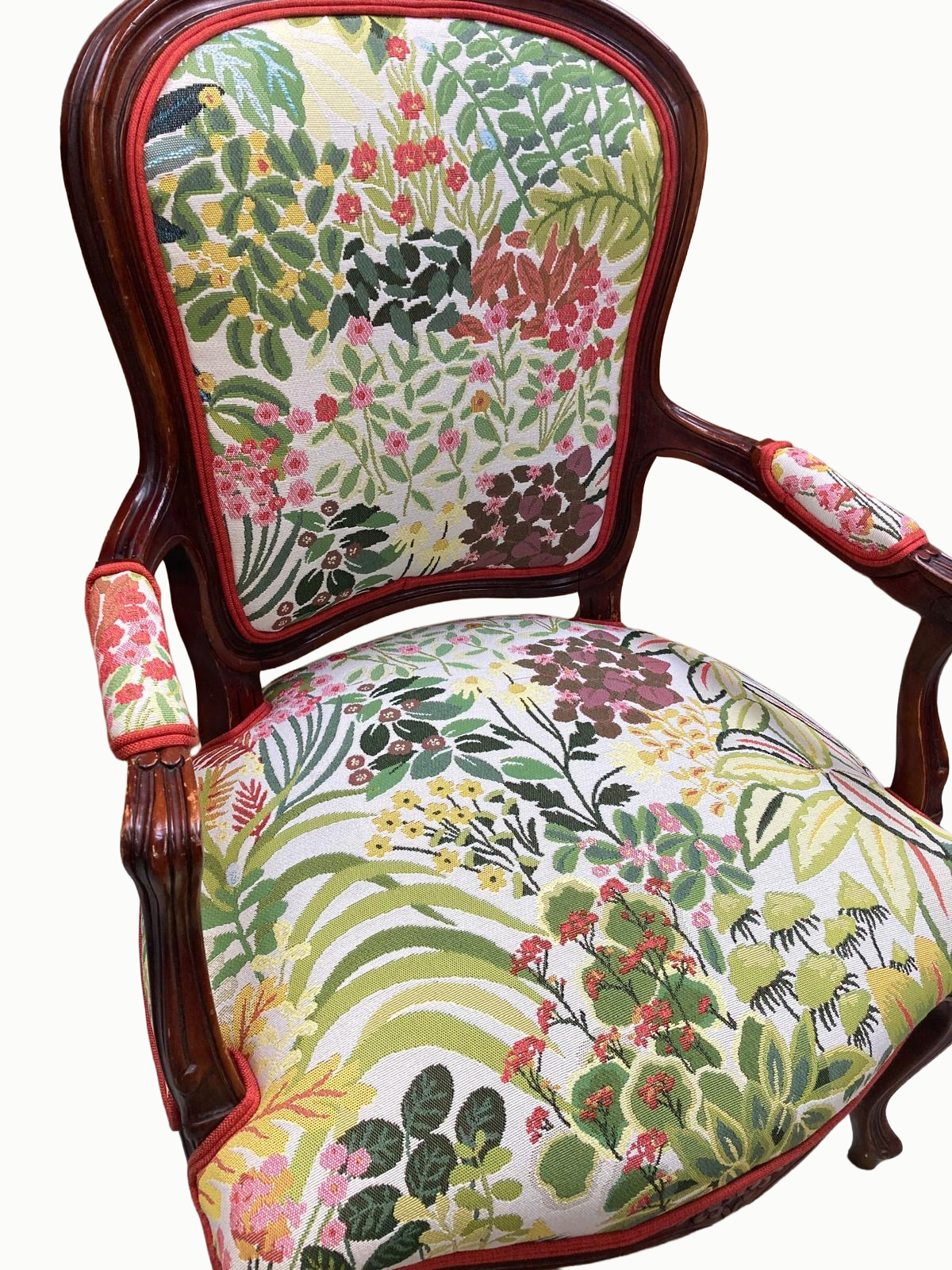 A vintage wooden armchair with a floral tapestry upholstery featuring various green leaves and colorful flowers, with red piping accents.