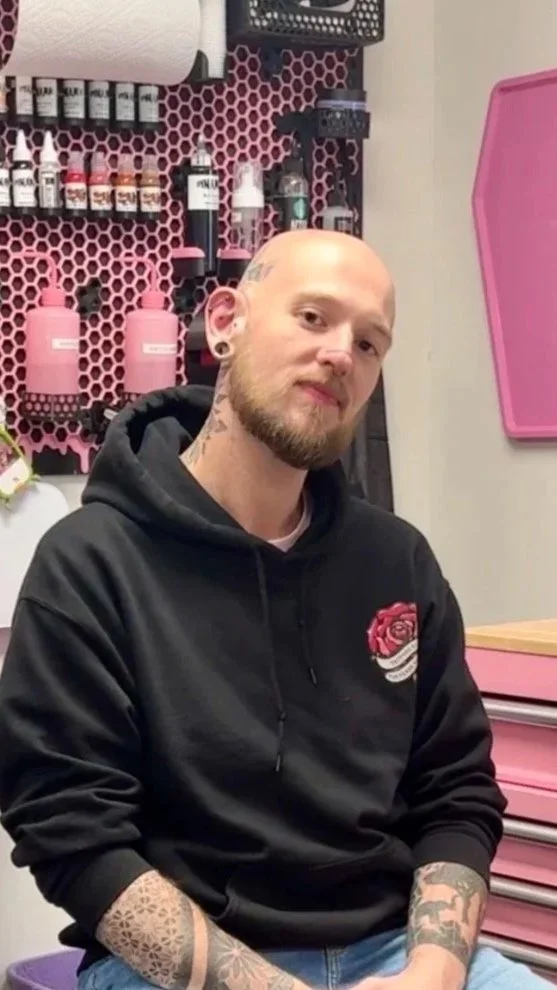 A man with a beard and tattoos on his arms and neck, wearing a black hoodie with a rose graphic, sitting in a tattoo parlor.