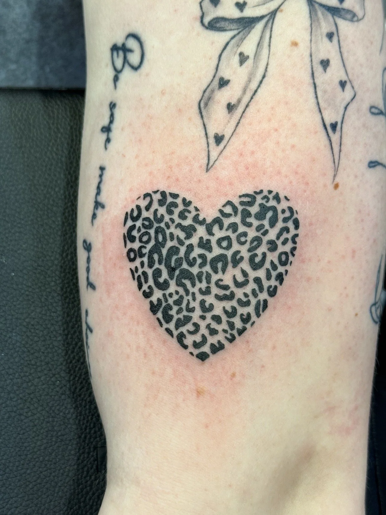 Tattoo of a leopard print heart with a small tattoo of a bow and a phrase along the side.