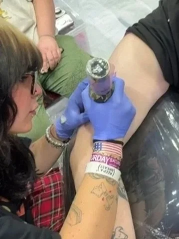 Person receiving a tattoo on their arm at a tattoo parlor, with a tattoo artist applying ink using a tattoo gun, while another person watches in the background.