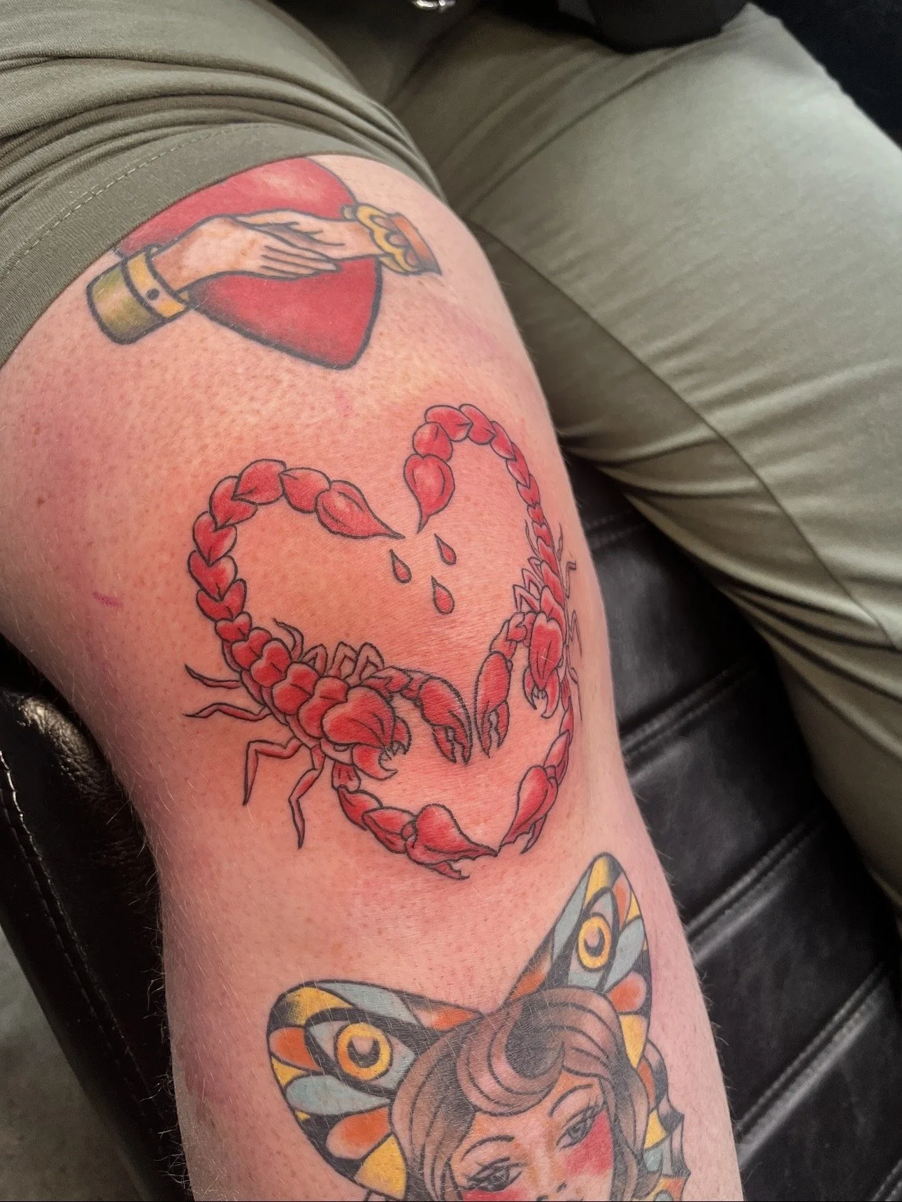 Tattoo of a red heart with a hand holding a yellow flower on top, and a red scorpion with a heart-shaped tail and a woman's face with a butterfly on her head below.