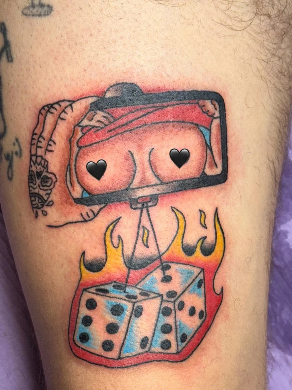Tattoo of a hand holding a phone, with a photo of a woman's buttocks inside the phone screen. The buttocks have black heart icons over the nipples. Below the phone, there are dice and flames.
