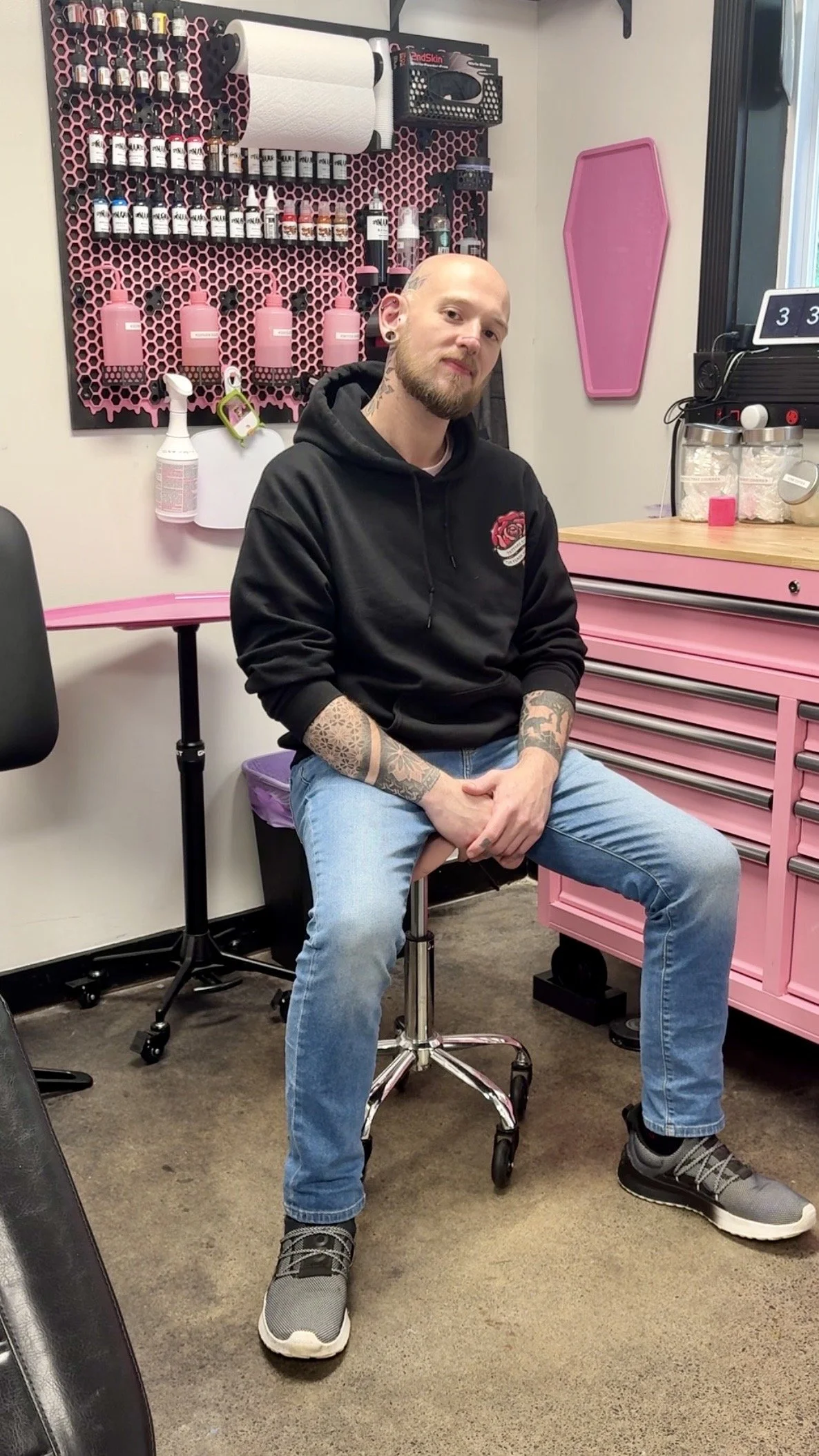 A man with a beard and tattoos sitting on a stool in a tattoo shop, surrounded by pink storage cabinets and tattoo supplies.