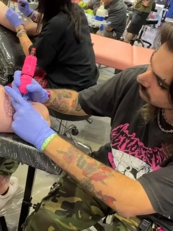 A tattoo artist wearing a black shirt with pink lettering is tattooing a person's arm in a busy tattoo shop. The artist has long hair, tattoos on both arms, and is wearing blue gloves. The client, seated on a chair, has their arm extended and is getting a colorful tattoo. Several people are working or sitting in the background.