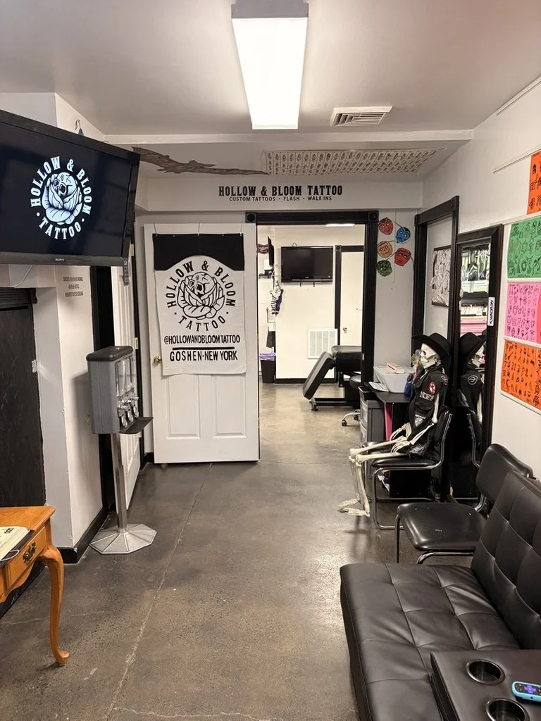 Interior of Hollow & Bloom Tattoo shop with a logo on a sign, black chairs, a TV, and colorful artwork on the wall.