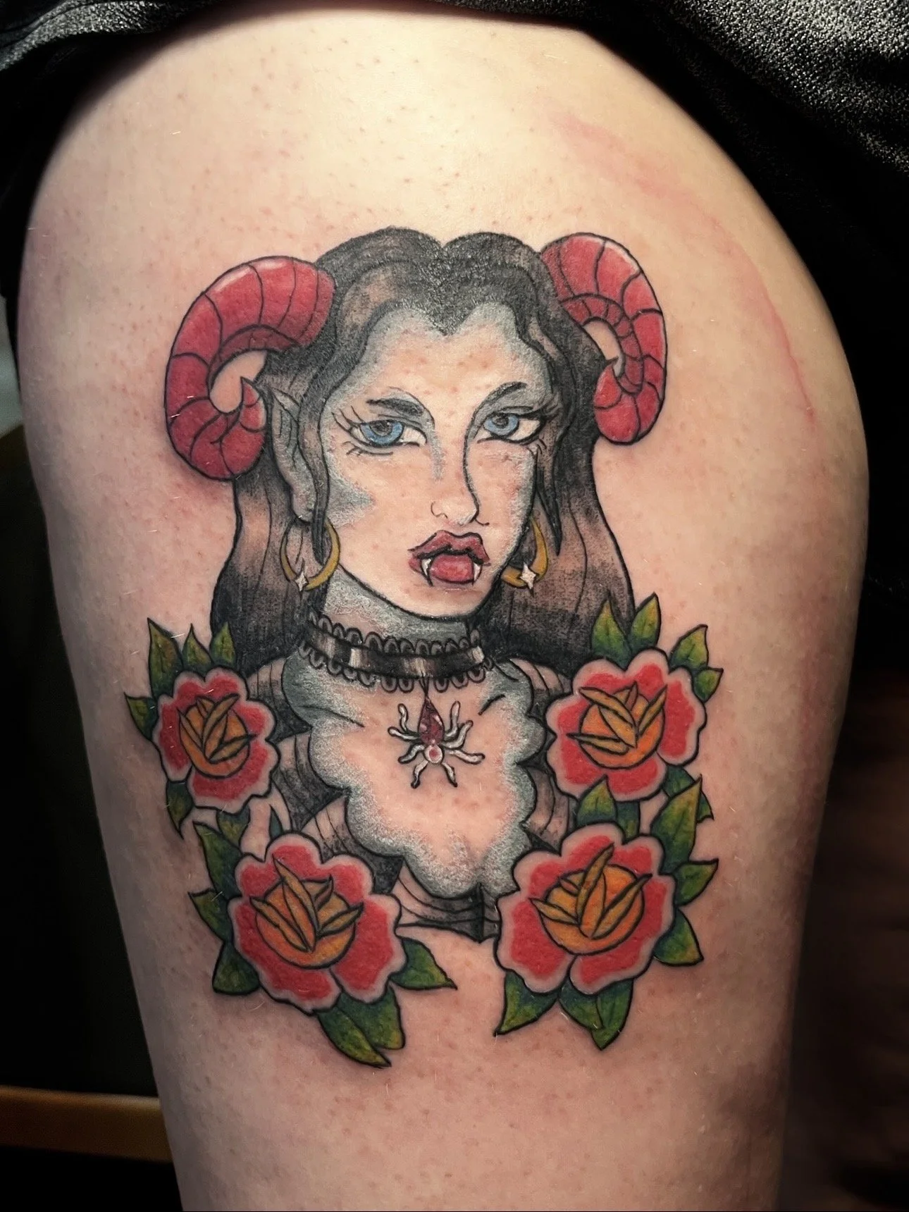 Colorful tattoo of a woman with black hair, blue eyes, red horns, vampire-like fangs, and floral and decorative jewelry, surrounded by red and orange flowers with green leaves.