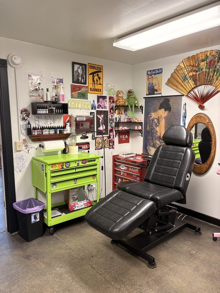Tattoo artist's workspace with a black tattoo chair, a green rolling cart with a paper towel roll, shelves with ink bottles, posters, a fan, a mirror, and various tattoo art decorations on the wall.