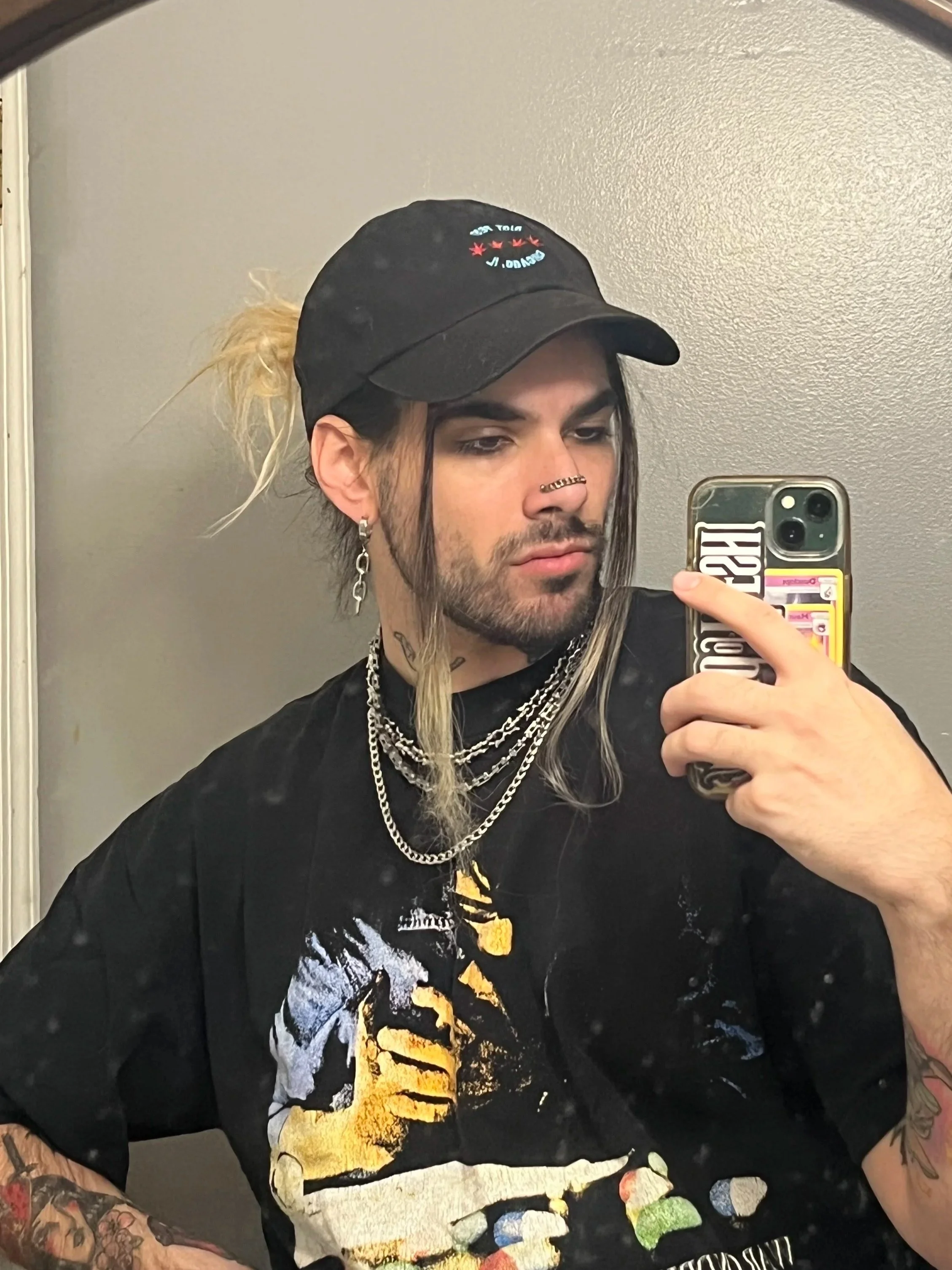 A person taking a mirror selfie with a smartphone, wearing a black cap, multiple necklaces, and a black T-shirt with a colorful graphic design. The person has tattoos on their arms, earrings, and a septum piercing. The background consists of a plain beige wall.