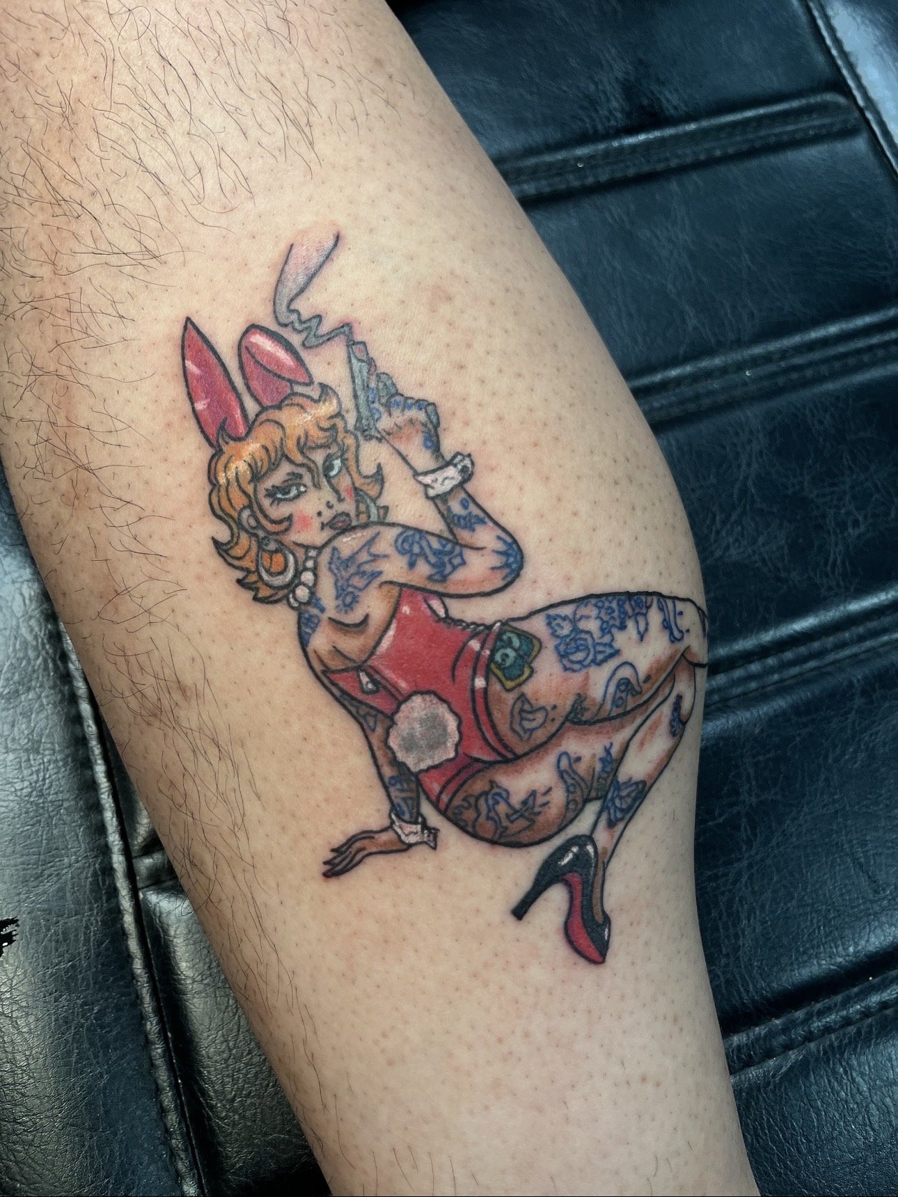 Tattoo of a stylized woman with blonde hair, wearing a red dress with floral tattoos on her arms and legs, holding a cigarette holder with a lit cigarette, and wearing pink bunny ears, sitting with one leg crossed over the other.